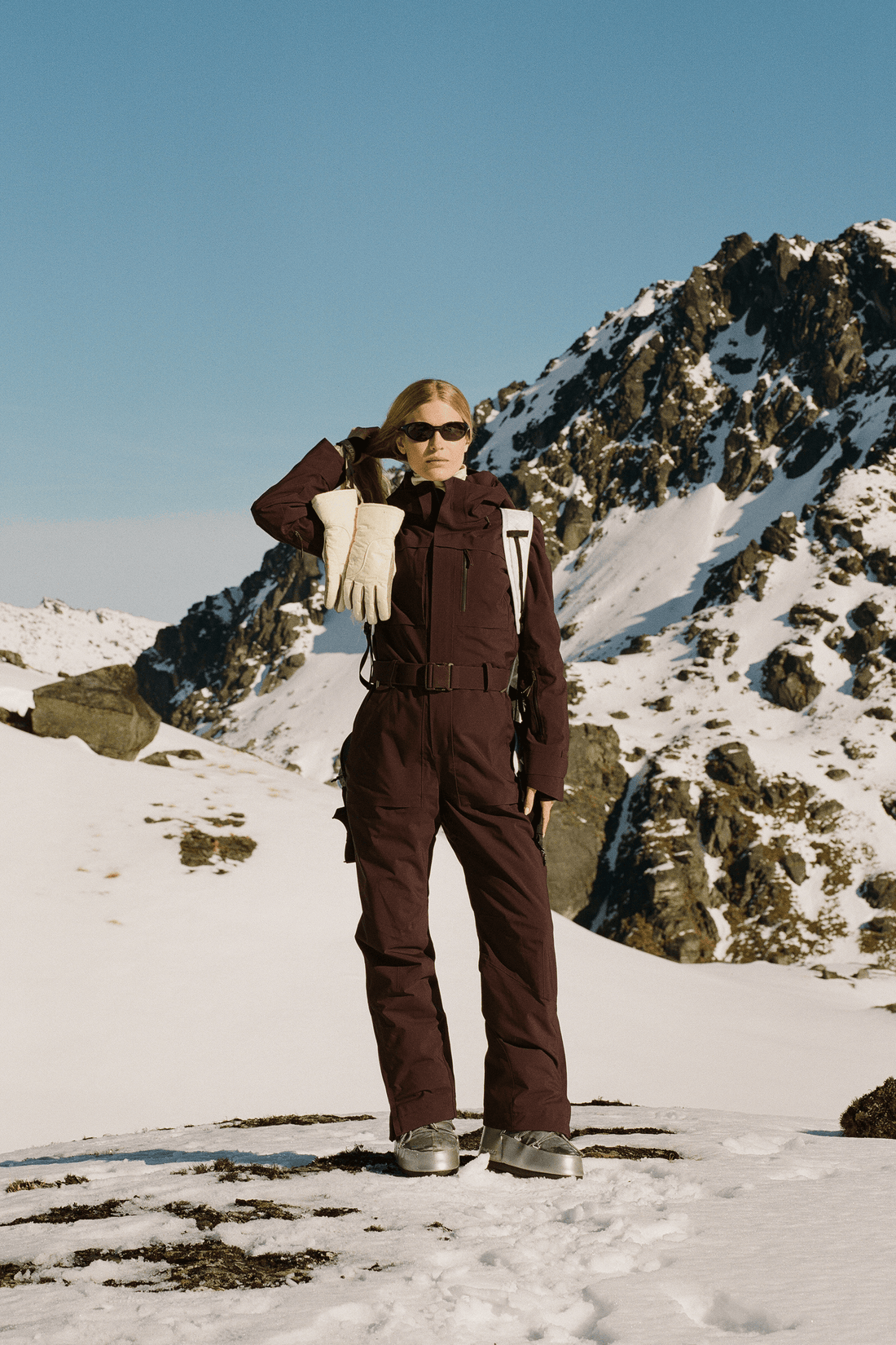 A model wearing a dark brown ski suit and sunglasses stands in a snowy mountain landscape under a clear blue sky, holding a white jacket.