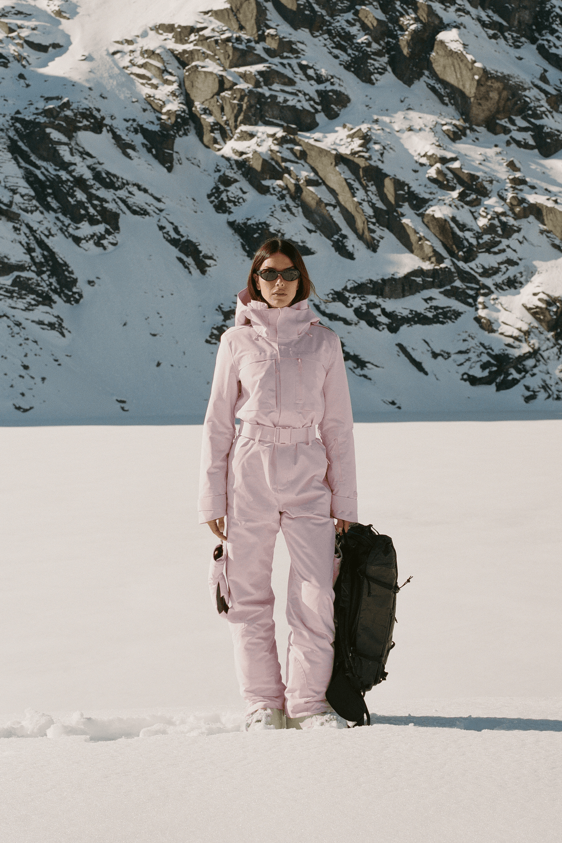 A woman wearing a light pink ski suit stands in the snow holding a black backpack, with a snowy mountain backdrop.