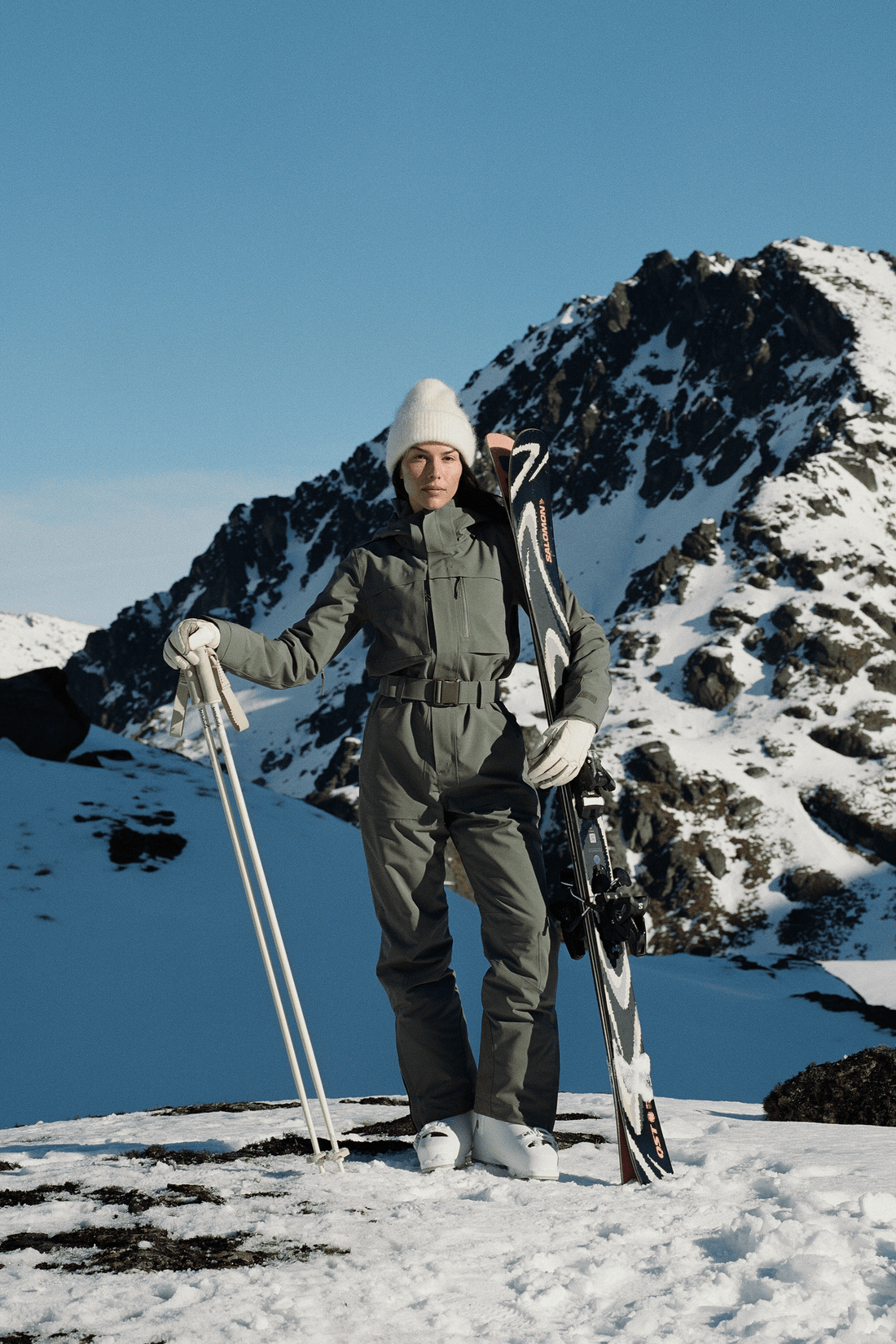 A person standing in snowy mountains wearing an olive green ski suit, white beanie, white gloves, and holding ski poles, with skis on their back against a clear blue sky.