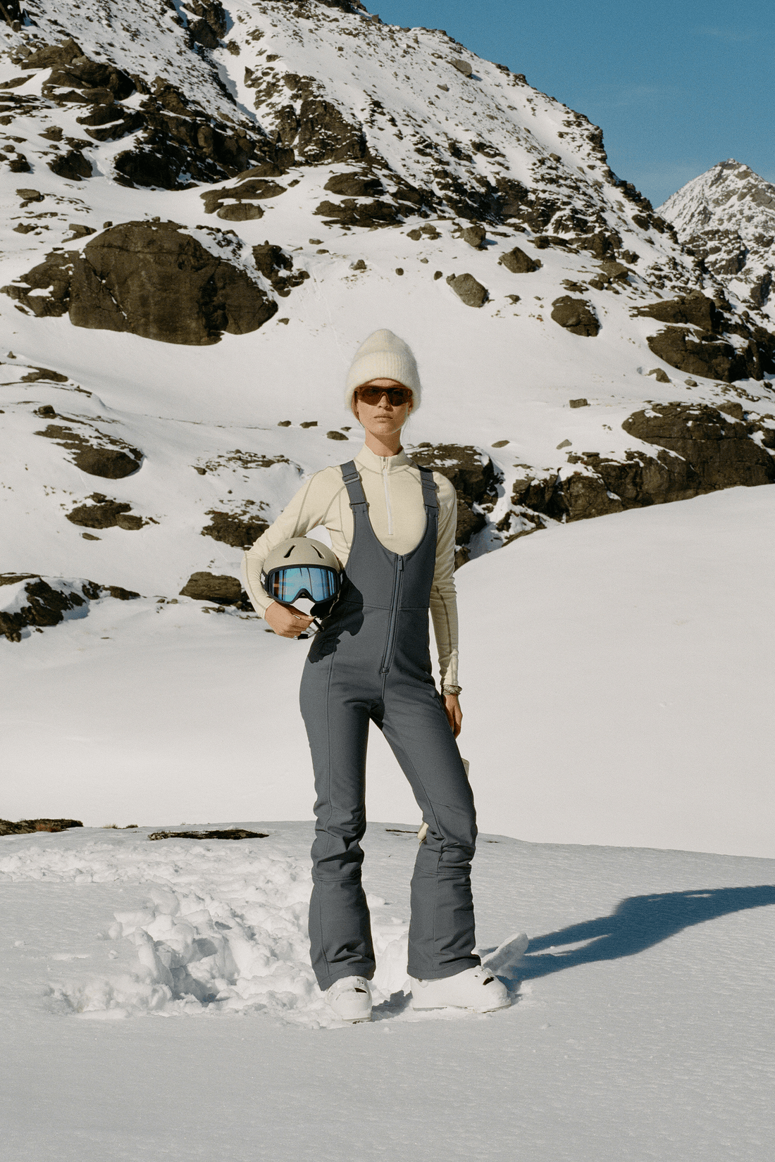 A person standing in a snowy mountain landscape wearing dark gray ski bib overalls, a cream long-sleeve top, a white beanie, and holding ski goggles.