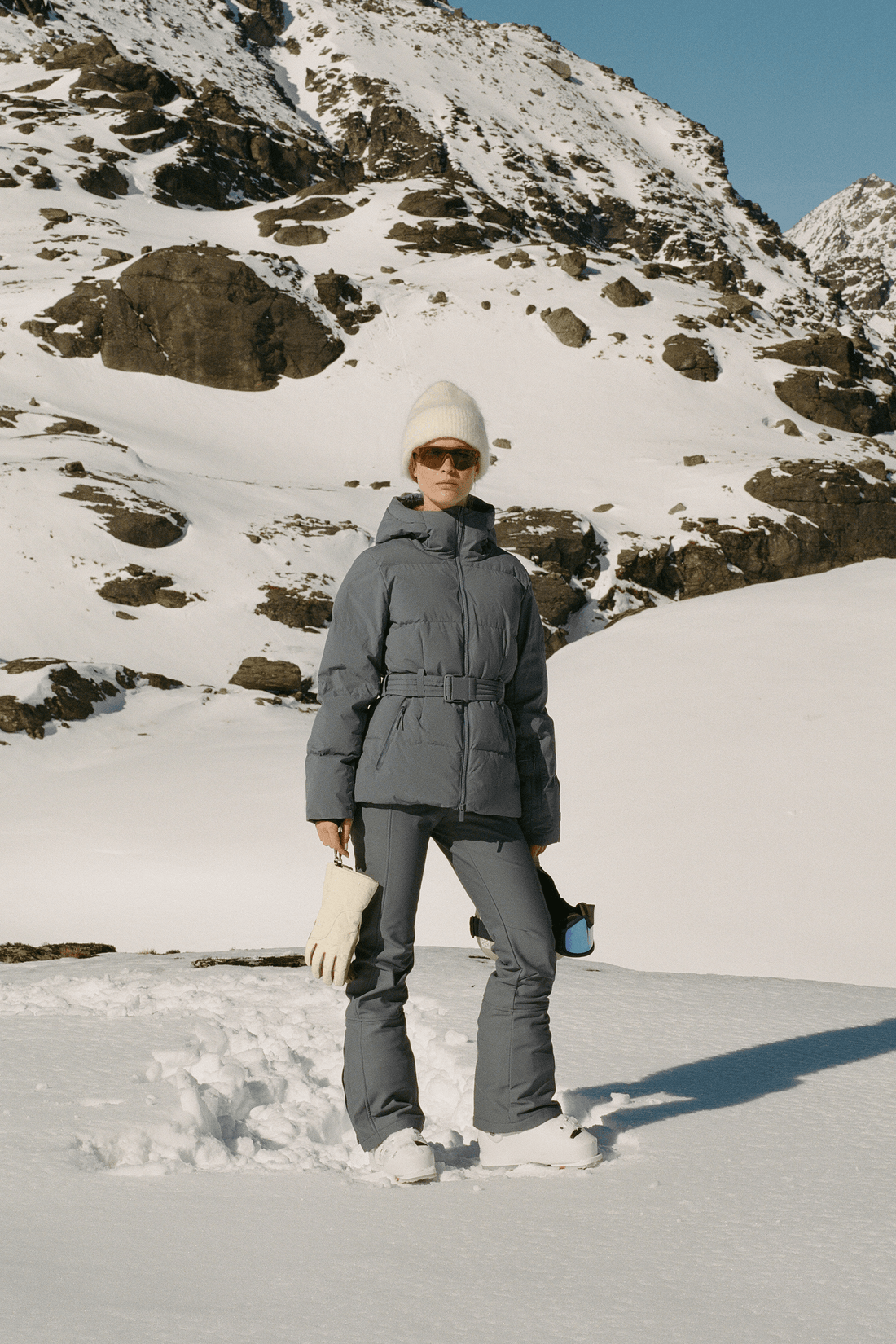 A person standing in the snow wearing a dark gray puffer jacket, gray ski pants, a white knit beanie, and white gloves, with rocky snowy mountains in the background.