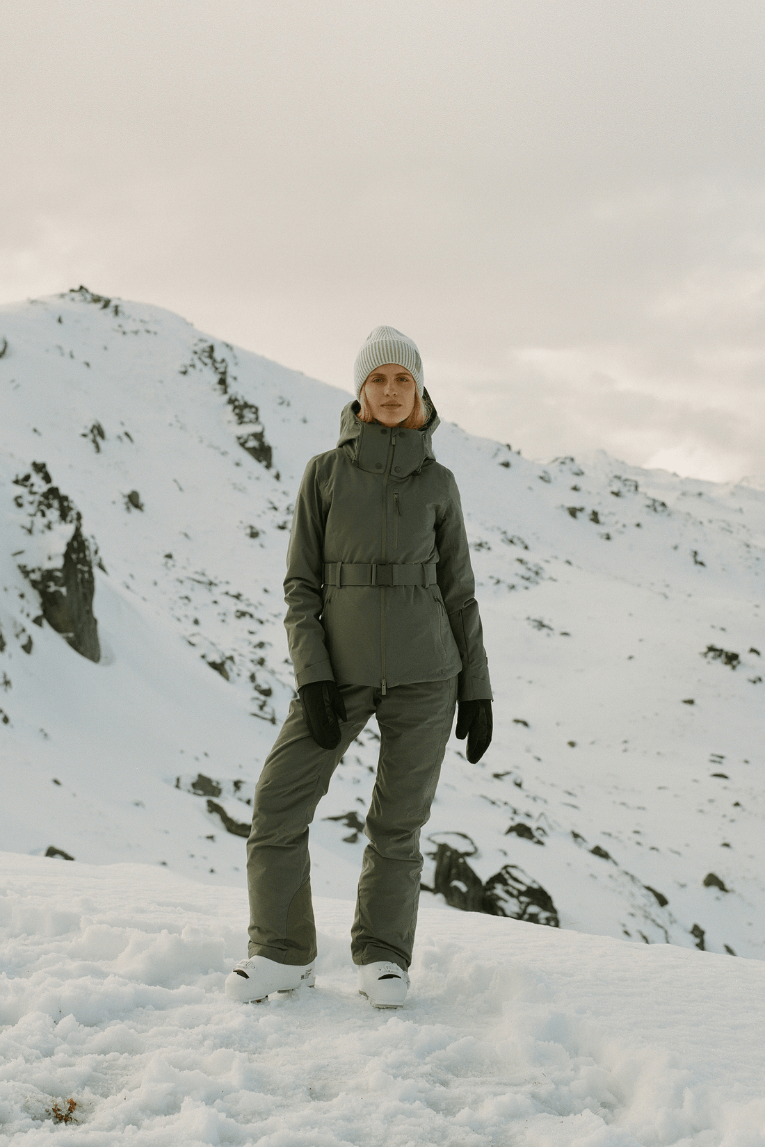 A person standing in the snow wearing an olive green ski jacket and matching pants, with a light gray beanie, black gloves, and white ski boots, against a snowy mountain background.
