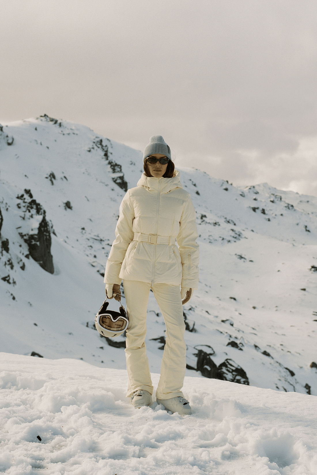 A model stands in a snowy mountain landscape wearing a cream-colored ski jacket and matching pants, holding ski goggles. The background features snow-covered peaks under a cloudy sky.