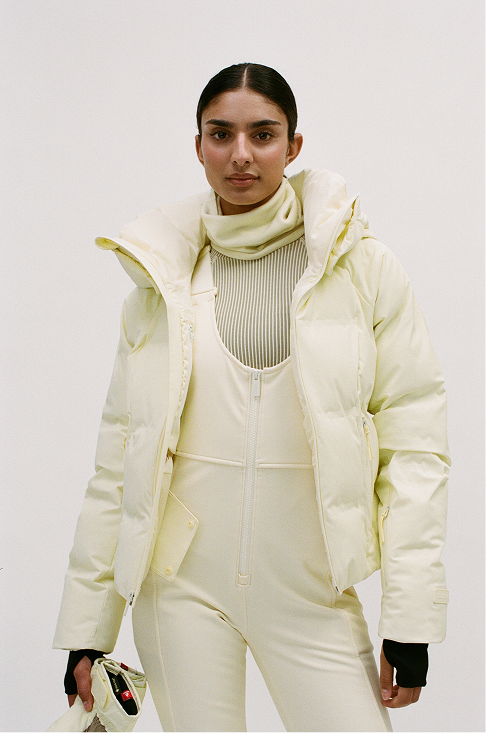 A woman from behind wearing a pale pastel yellow puffer jacket and matching light pants against a clean white studio background.