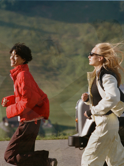 Two people running outdoors near a vehicle, one wearing a bright red puffer jacket and dark pants, and the other in a light beige and black jacket with white pants. The background shows a blurred green landscape.