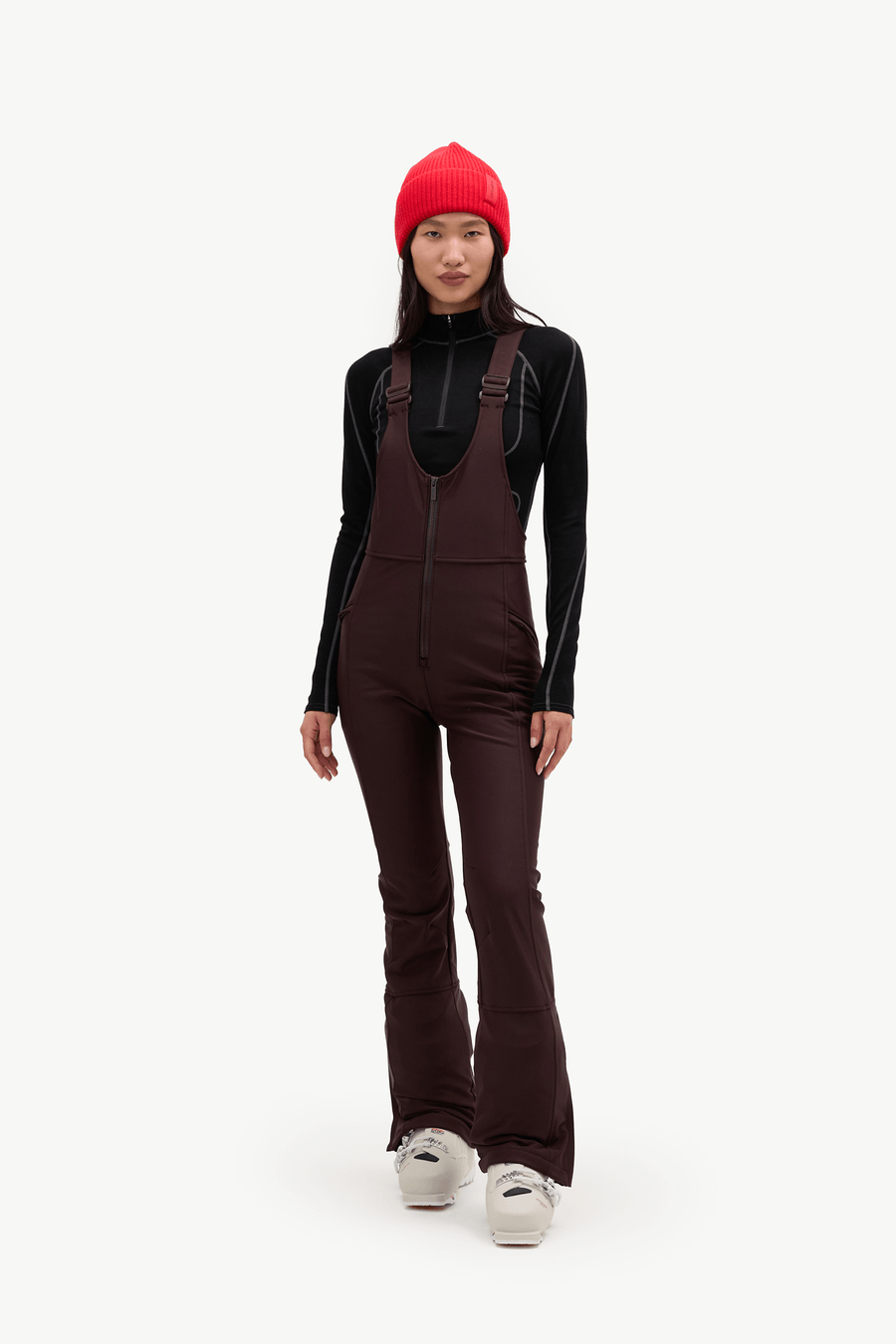 A model wearing a dark brown ski bib overalls, a black long-sleeve top, a bright red beanie, and white sneakers, standing against a solid white studio background.