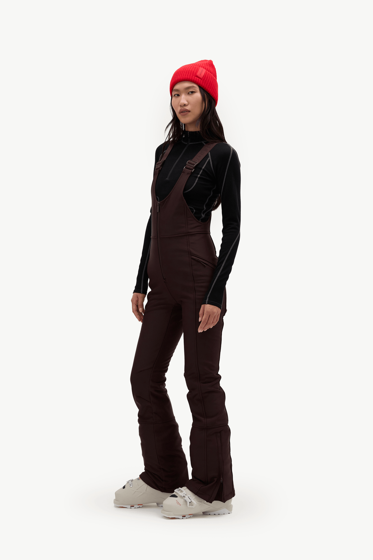 A model wearing a dark brown ski jumpsuit over a black long-sleeve top, a bright red beanie, and off-white Croc sneakers, standing against a plain white studio background.