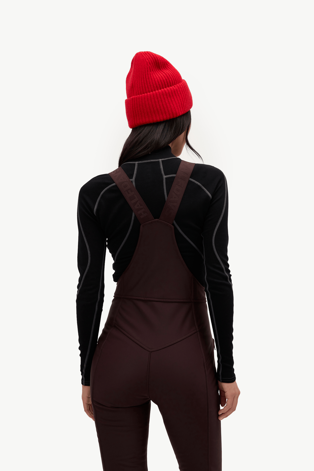 A model shown from behind wearing a bright red knit beanie, a black long-sleeve athletic top, and dark brown bib overalls against a plain white background.