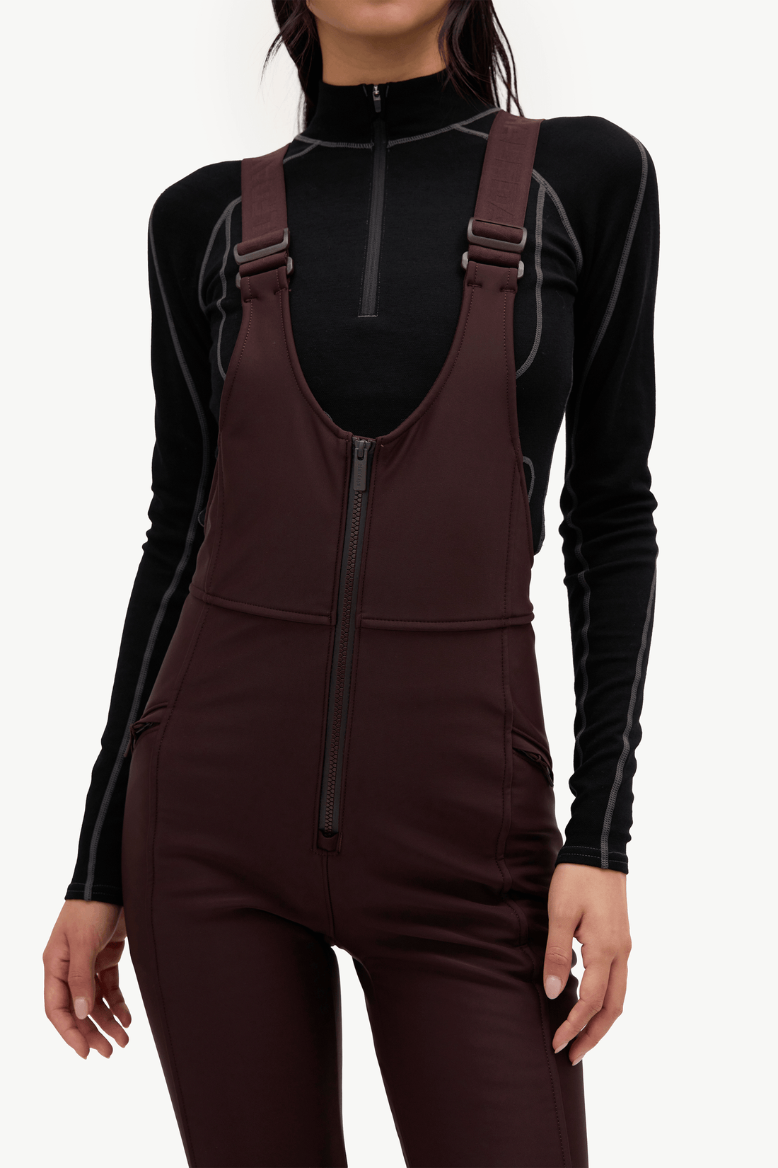 A model wearing dark brown snow bib overalls, a black long-sleeve top, and a bright red knit beanie, posed against a solid off-white background.