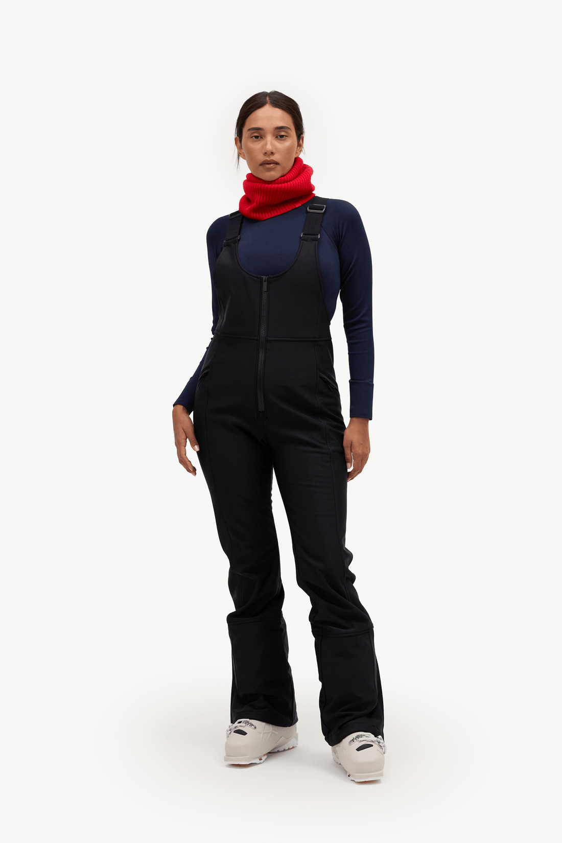 A model stands against a plain white studio background wearing a dark navy and black ski bib overalls with a red neck gaiter and light gray ski boots.