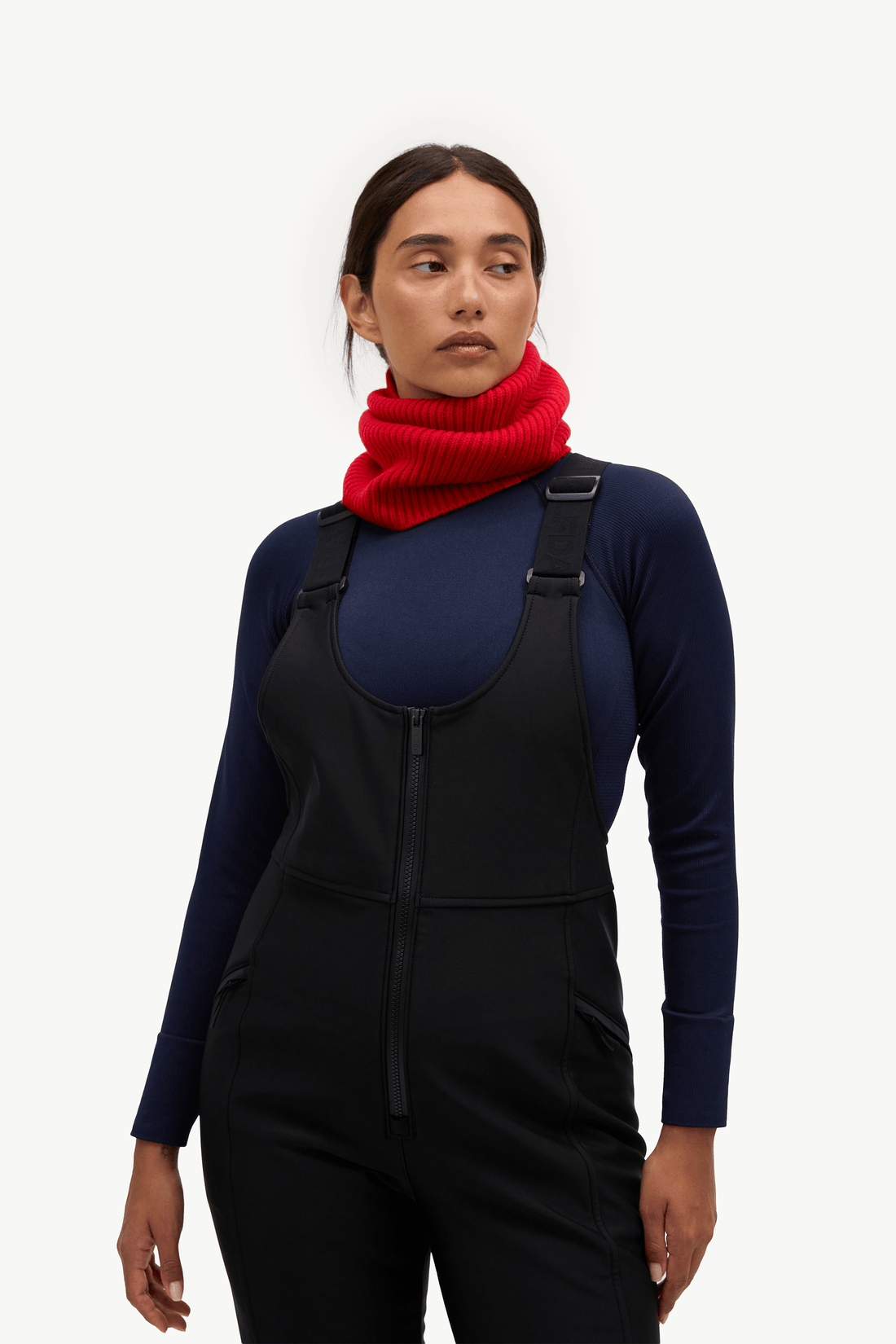 A woman modeling a black bib snowsuit over a navy blue long-sleeve top, accessorized with a bright red knit neck warmer, against a plain white studio background.