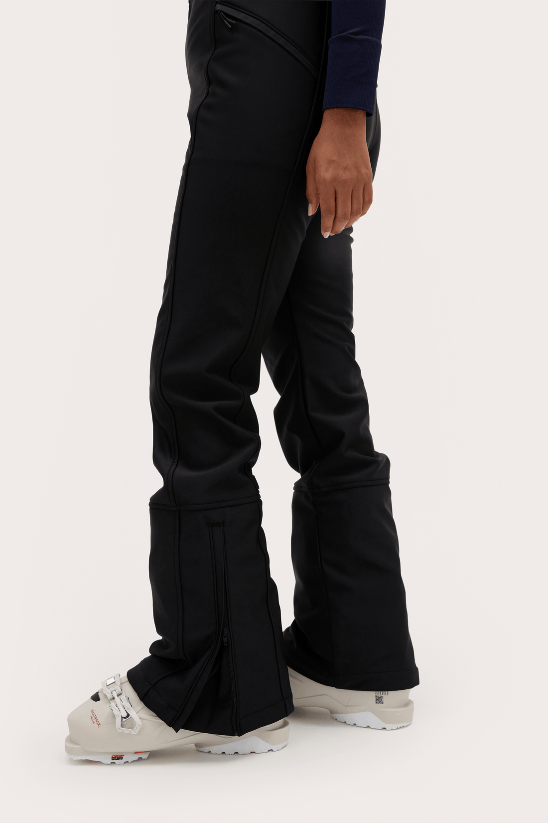 Black flared ski pants worn with ski boots, shown from the waist down on a light gray studio background.