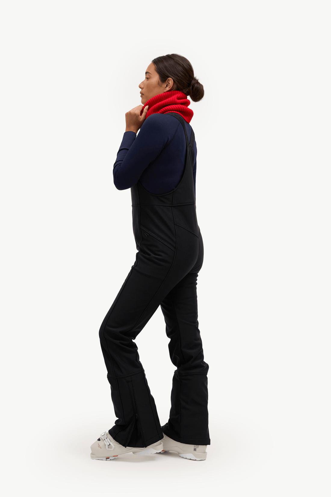 A woman in profile wearing a dark navy long-sleeve top, black snow pants, and a bright red knit neck warmer, standing against a solid white studio background.