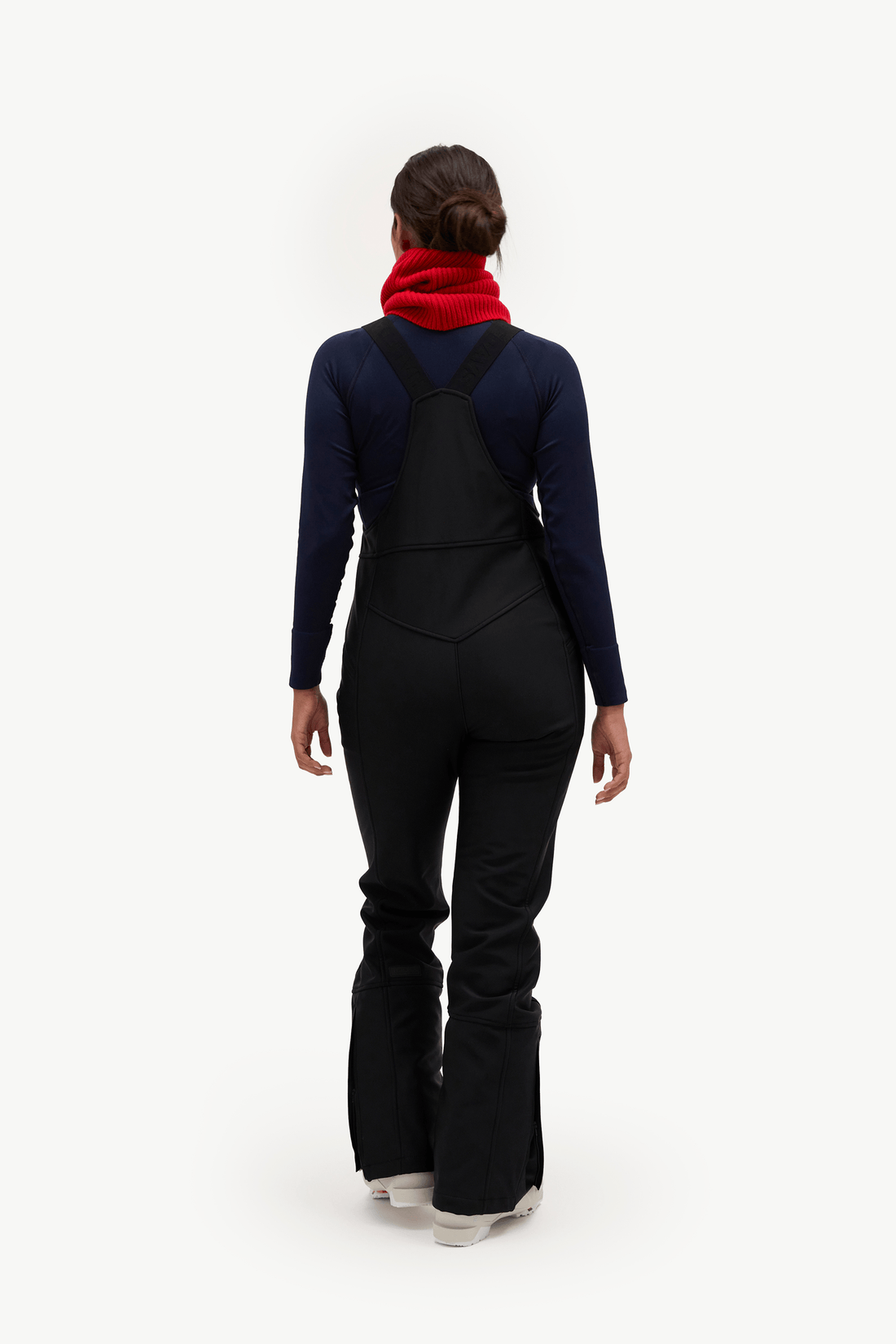 A model viewed from the back wearing a dark navy long-sleeve top, black bib-style overalls, a bright red neck warmer, and white sneakers against a plain white studio background.