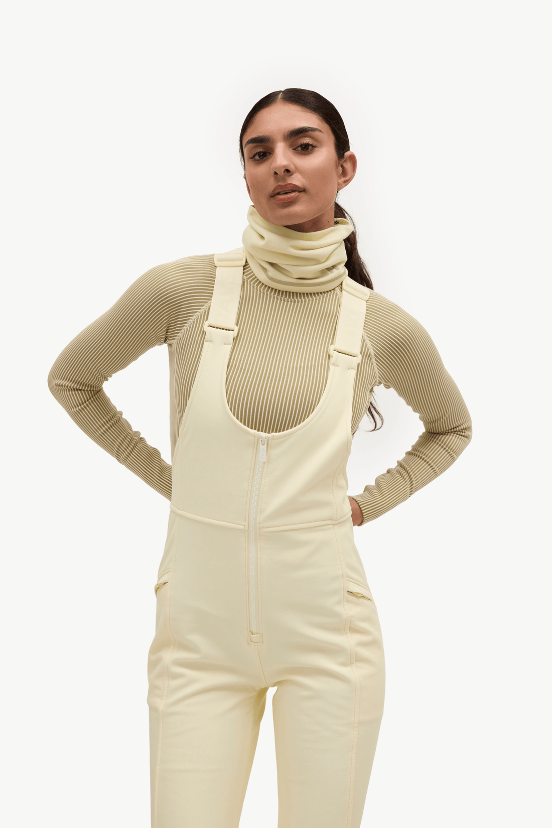 A model wearing cream-colored snow bib overalls over a ribbed beige turtleneck top, posed against a solid off-white studio background.