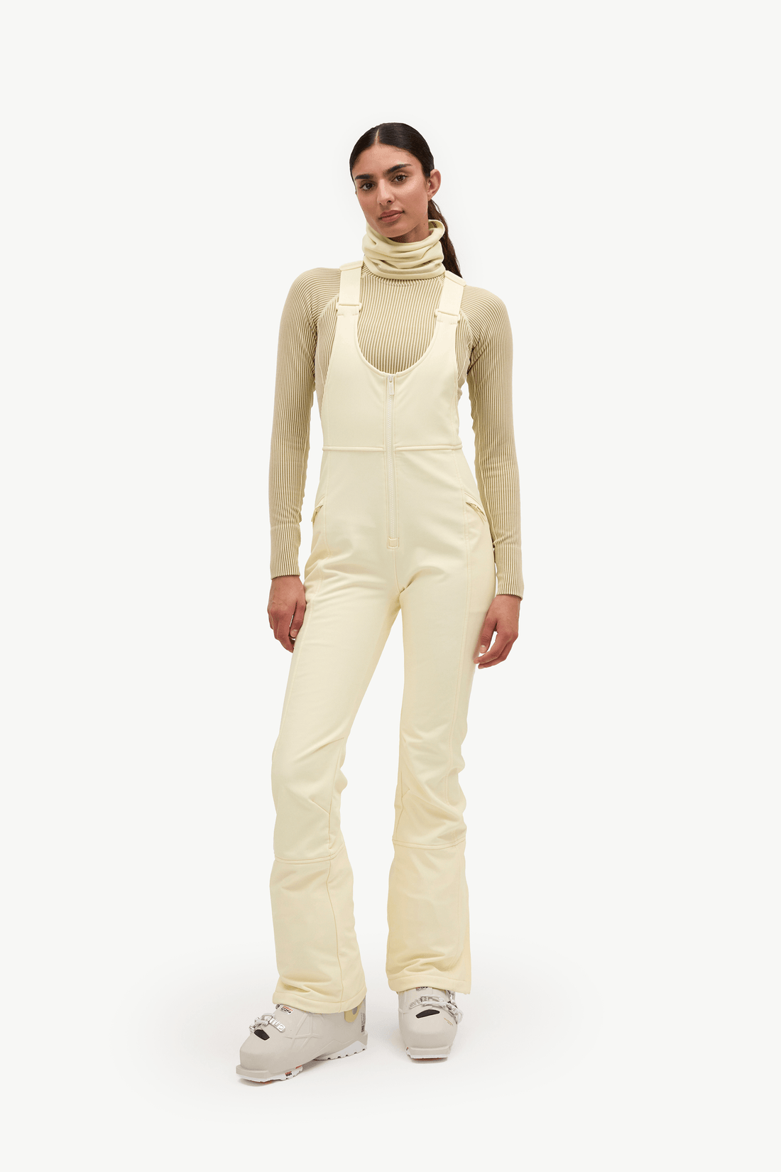 A model stands against a plain white studio background wearing cream-colored snow bib overalls over a beige long-sleeve turtleneck top and light-colored ski boots.