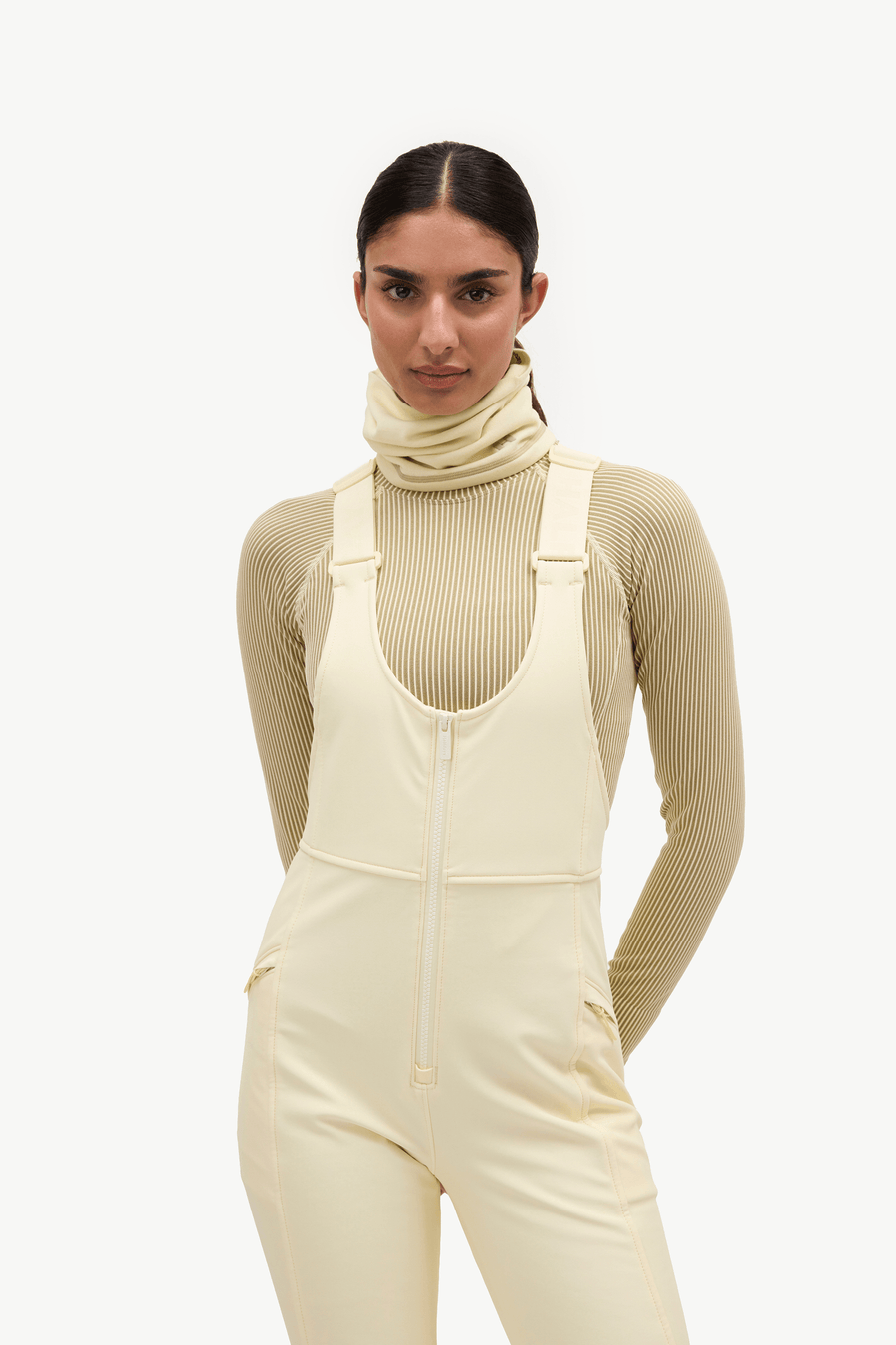 A model wearing a cream-colored ski jumpsuit over a matching ribbed knit turtleneck sweater, photographed against a solid off-white studio background.