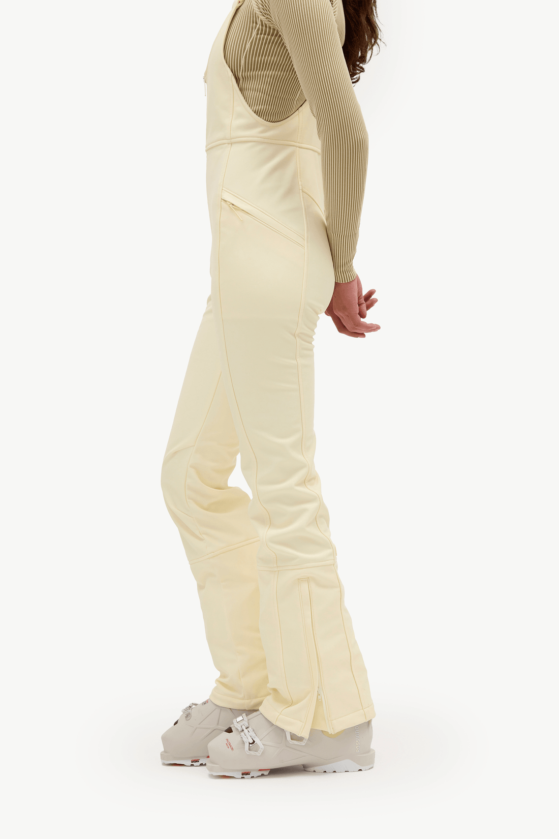 A model shown from the side wearing cream-colored snow bib overalls with integrated pockets, a beige ribbed long-sleeve top, and light gray ski boots, set against a plain white studio background.