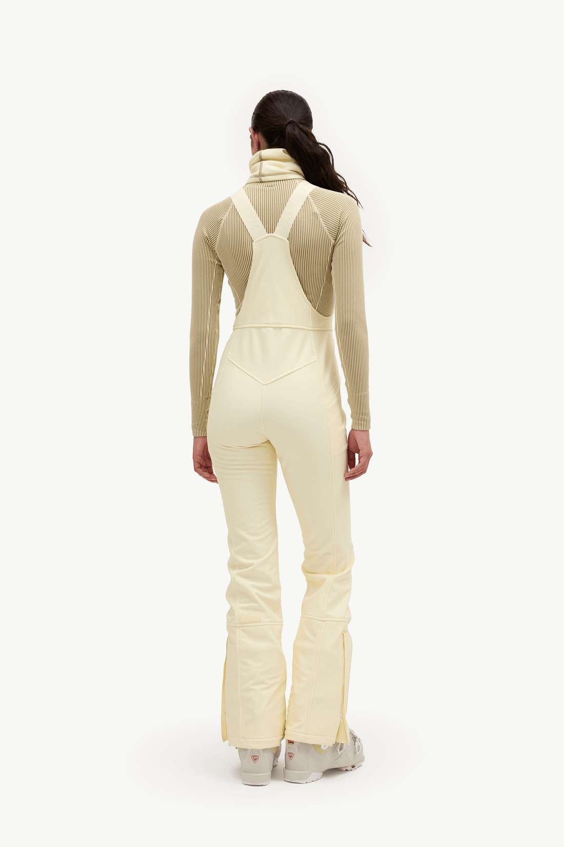 A model viewed from the back wearing a light cream ski jumpsuit with integrated shoulder straps over a beige long-sleeve top, set against a plain white studio background.