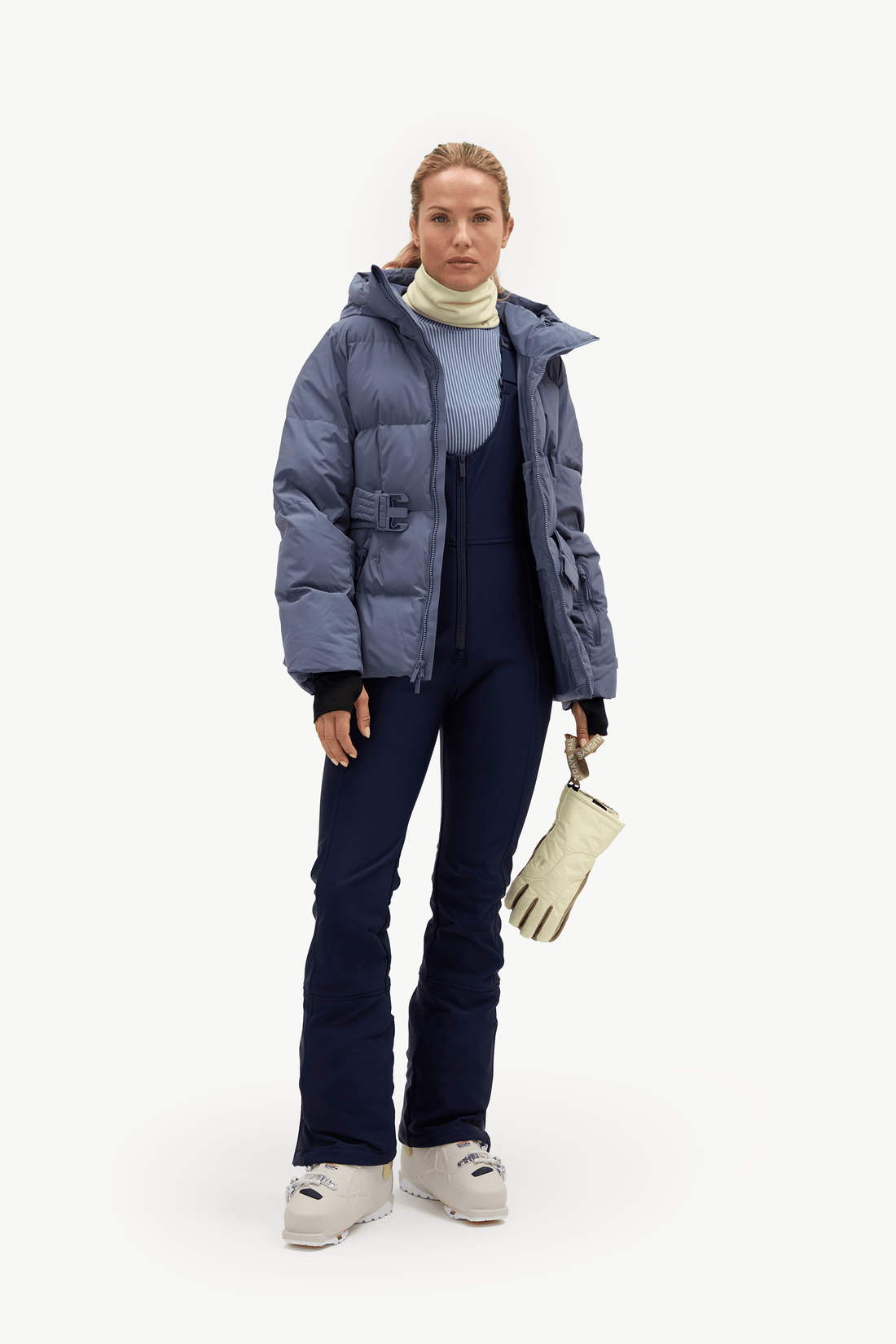 A woman stands against a plain white studio background wearing a blue puffer jacket over a light blue turtleneck, dark navy ski pants, white ski boots, and holding a cream-colored glove.