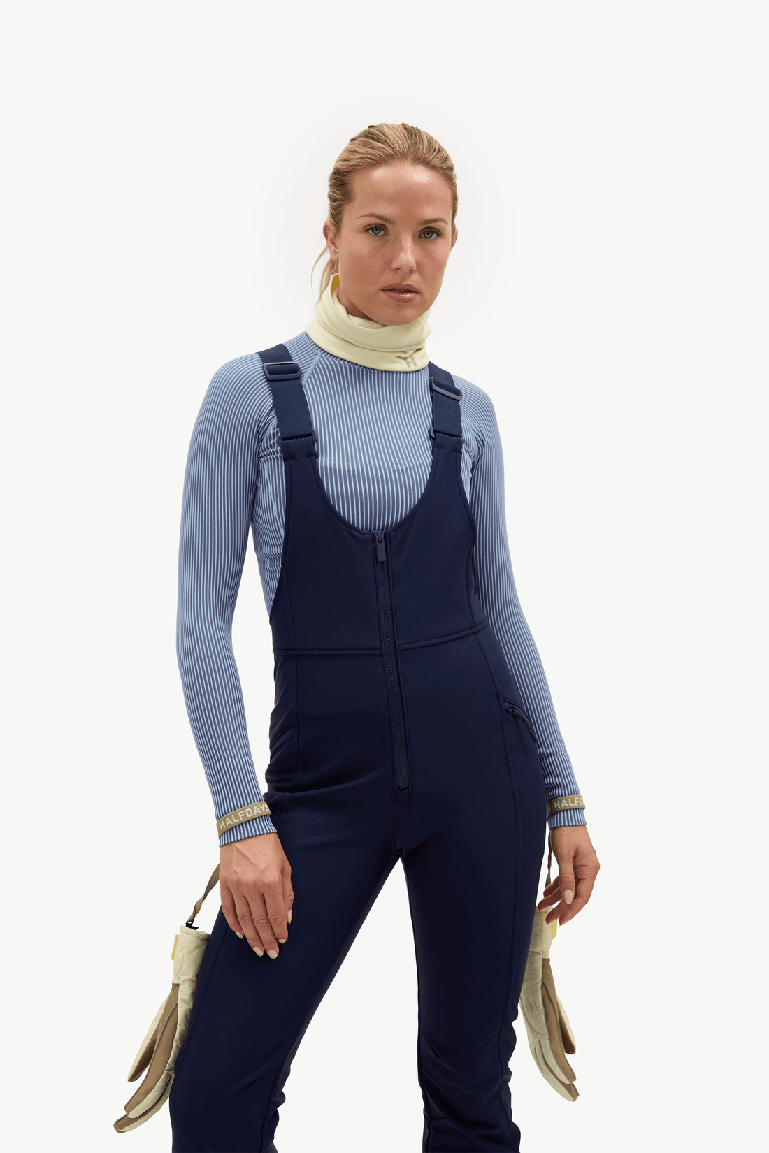 A woman modeling navy blue ski overalls over a light blue ribbed long-sleeve top and cream neck warmer, holding a beige handbag against a plain white studio background.