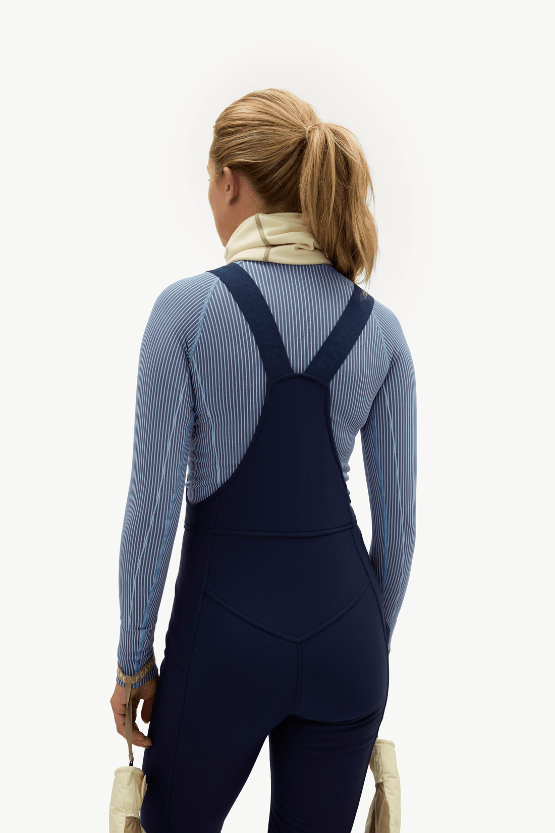 A woman shown from behind wearing a navy blue one-piece athletic jumpsuit over a light blue ribbed long-sleeve top, against a plain white studio background.