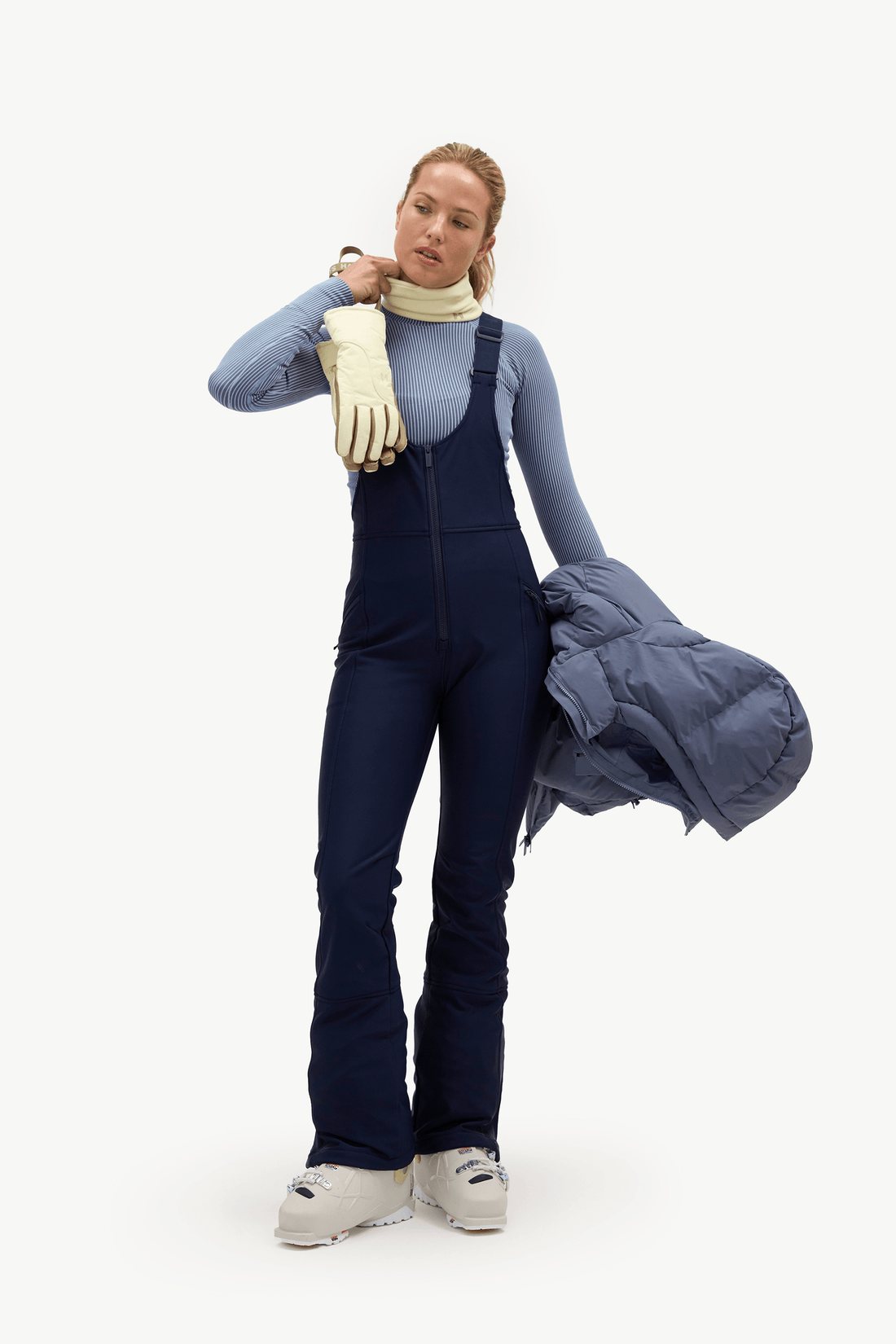 A woman modeling dark navy ski overalls with a light blue long-sleeve top, cream turtleneck, cream gloves, and white sneakers, while holding a gray puffer jacket against a plain white studio background.
