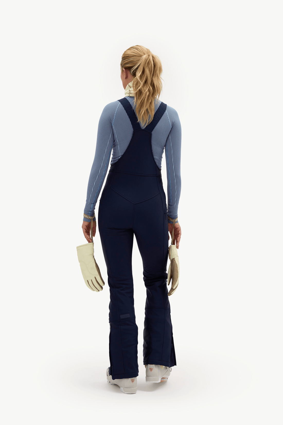 A woman viewed from behind wearing a dark navy blue ski jumpsuit over a dusty blue long-sleeve top, holding cream-colored gloves against a solid white studio background.