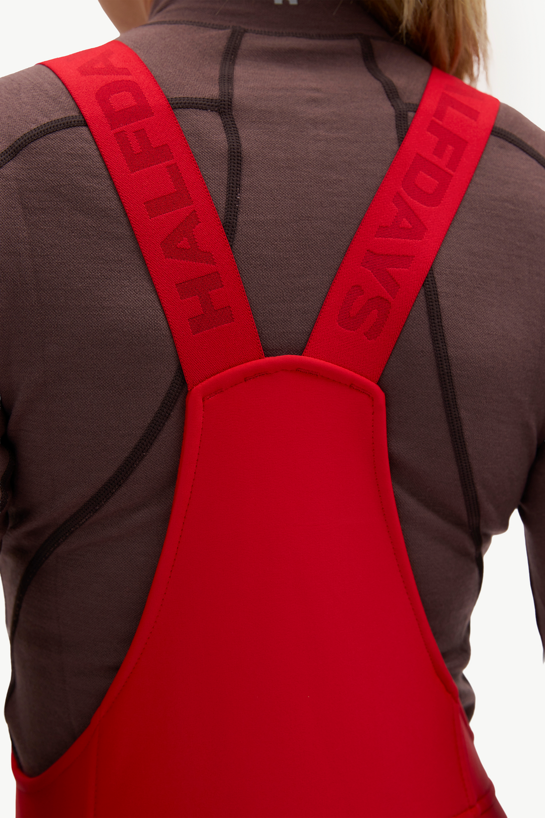 Close-up back view of a person wearing bright red bib overalls or snow pants over a dark brown athletic top. The red straps have the brand name HALFDAYS printed in dark red.