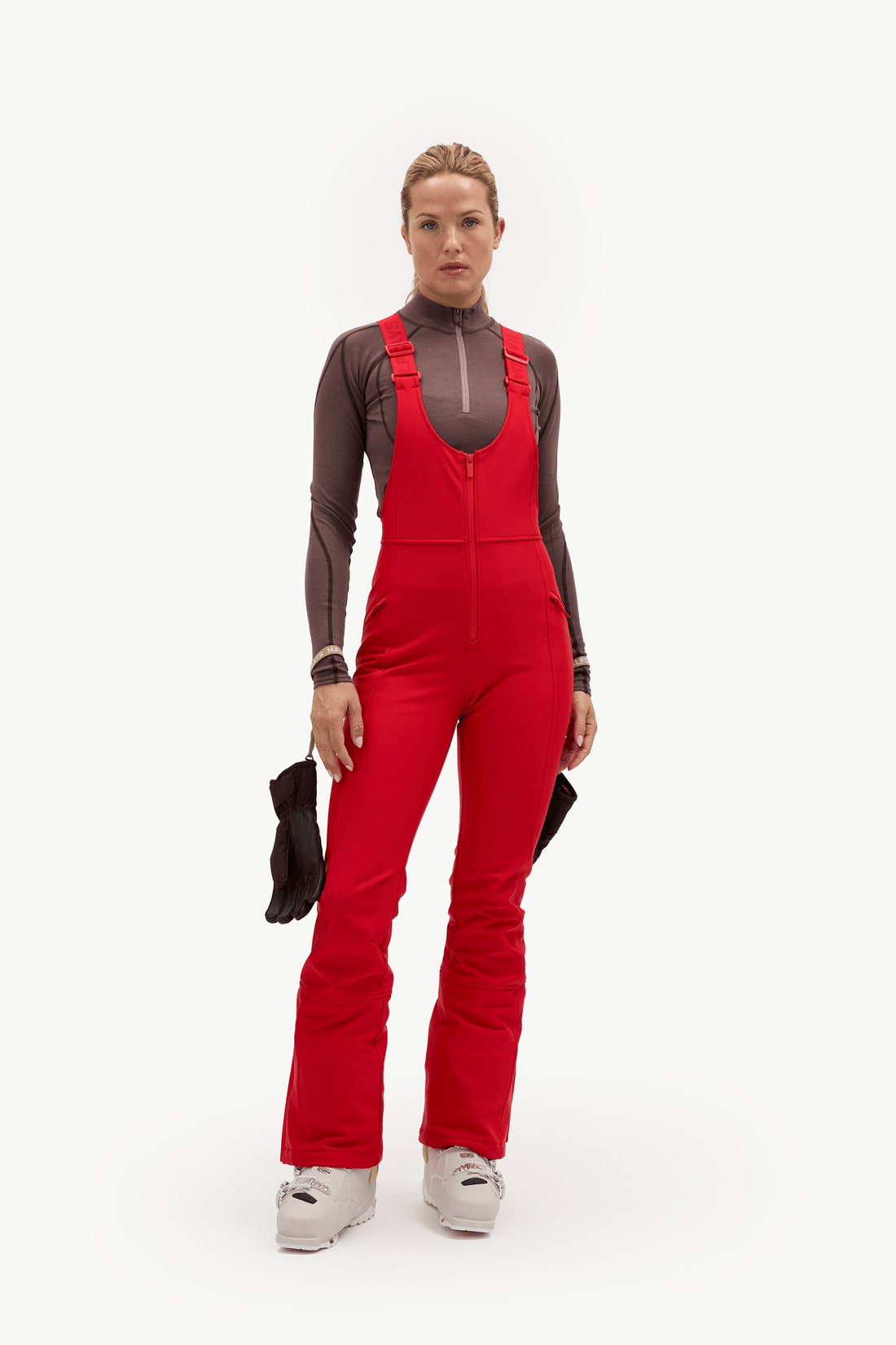 A model stands against a plain white studio background wearing a bright red ski bib over a dark brown long-sleeve top and white ski boots, holding a black glove in one hand.