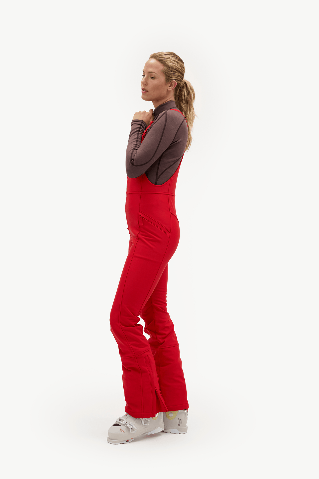 A woman modeling a bright red ski jumpsuit with a dark brown long-sleeve top underneath, standing in profile against a clean white studio background.
