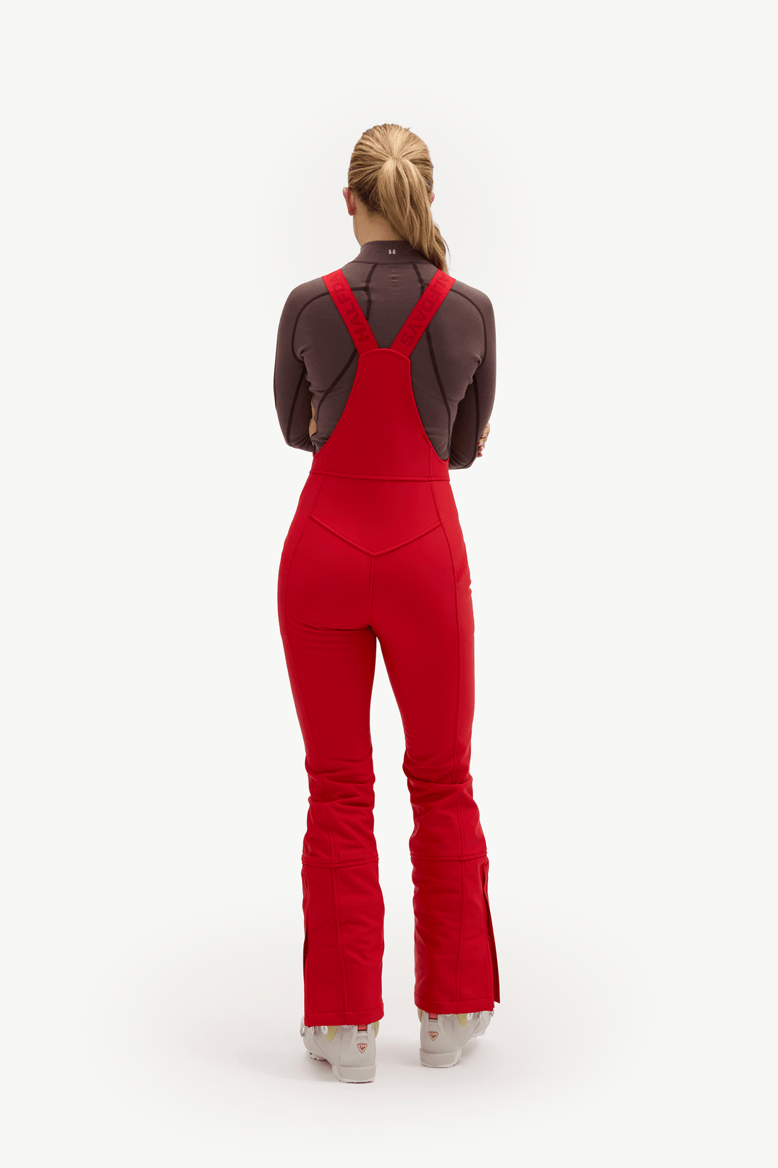 A woman viewed from behind wearing bright red bib overalls with a dark brown long-sleeve top and white sneakers, standing against a plain light gray studio background.