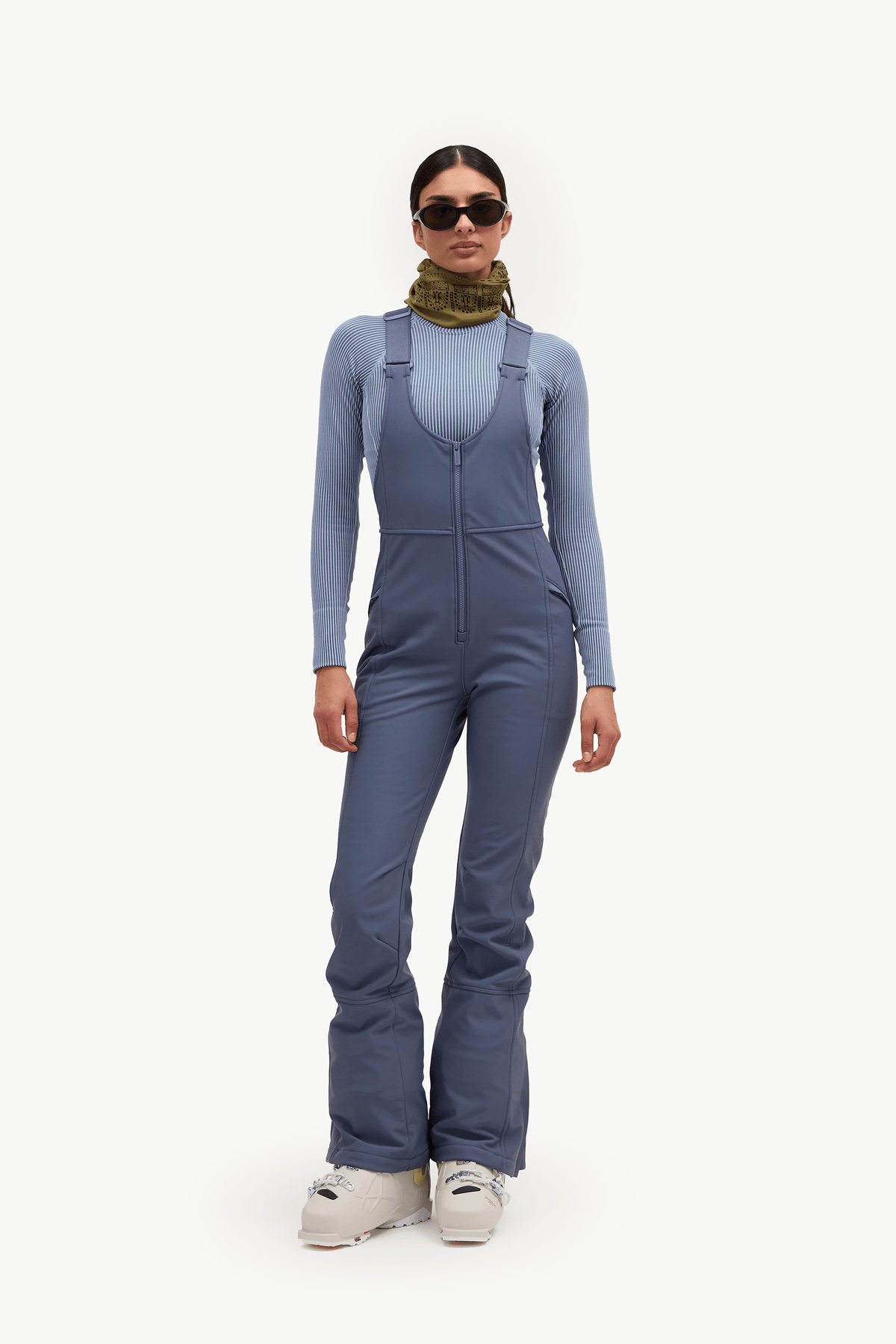 A model wearing slate blue ski bib overalls over a light blue long-sleeve top, olive green neck gaiter, and white ski boots, standing against a solid white background.