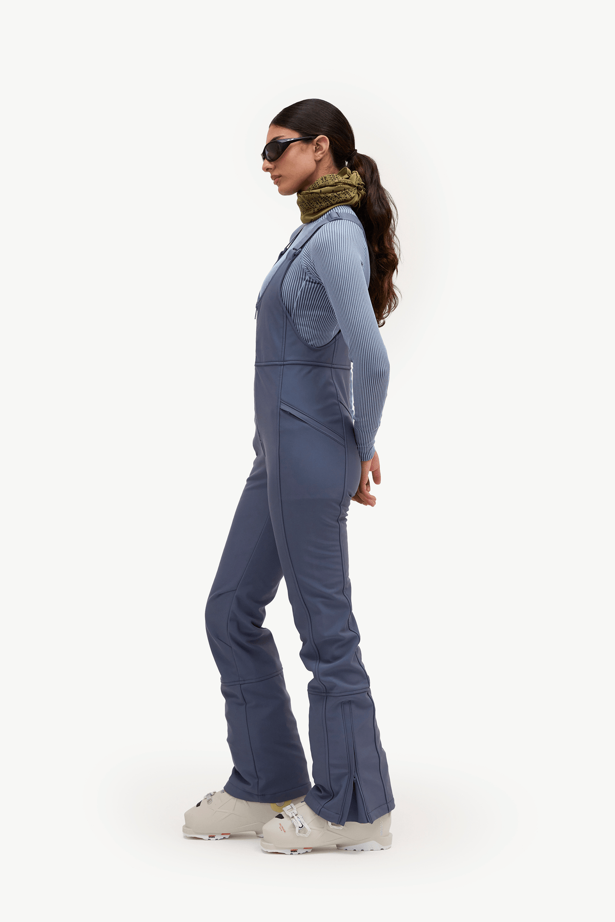 A woman in side profile wearing a slate blue ski jumpsuit, a green patterned neck warmer, sunglasses, and white ski boots against a plain white studio background.
