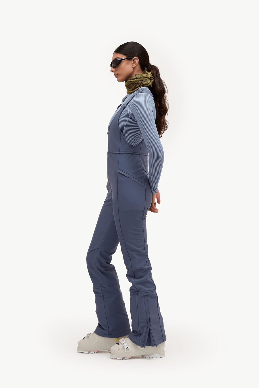 A woman in side profile wearing a slate blue ski jumpsuit, a green patterned neck warmer, sunglasses, and white ski boots against a plain white studio background.