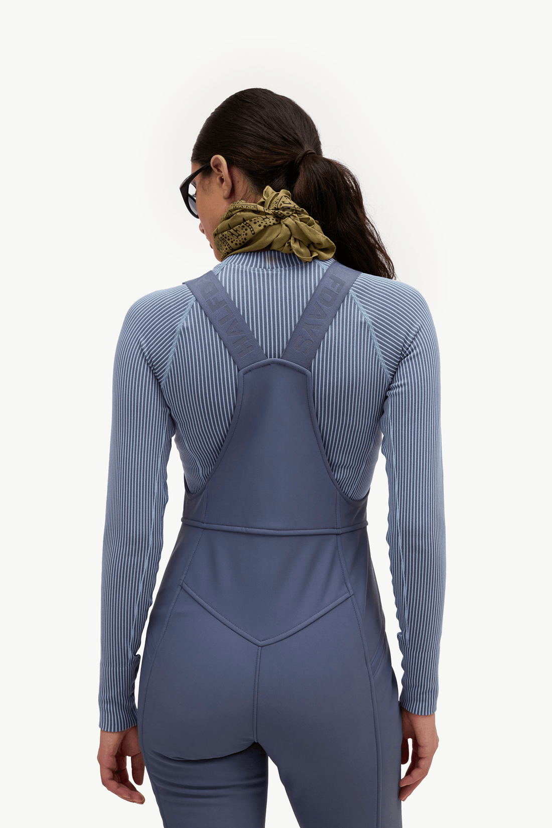 Rear view of a woman wearing a ribbed long-sleeve athletic jumpsuit in muted blue-gray with darker gray shoulder straps, against a plain light background.