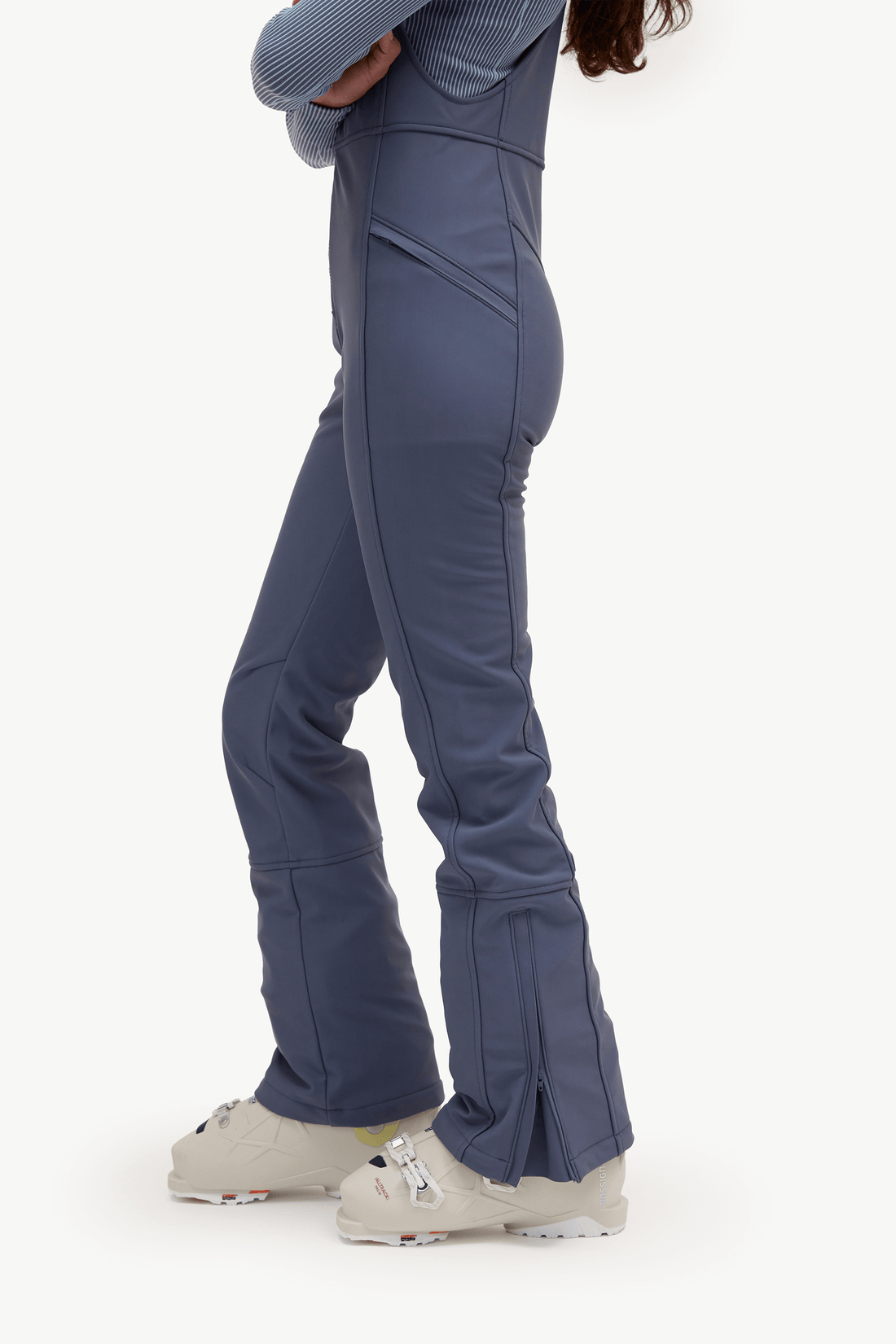 A side profile view of a person wearing slate blue ski pants with flared legs and light beige ski boots, set against a plain white studio background.