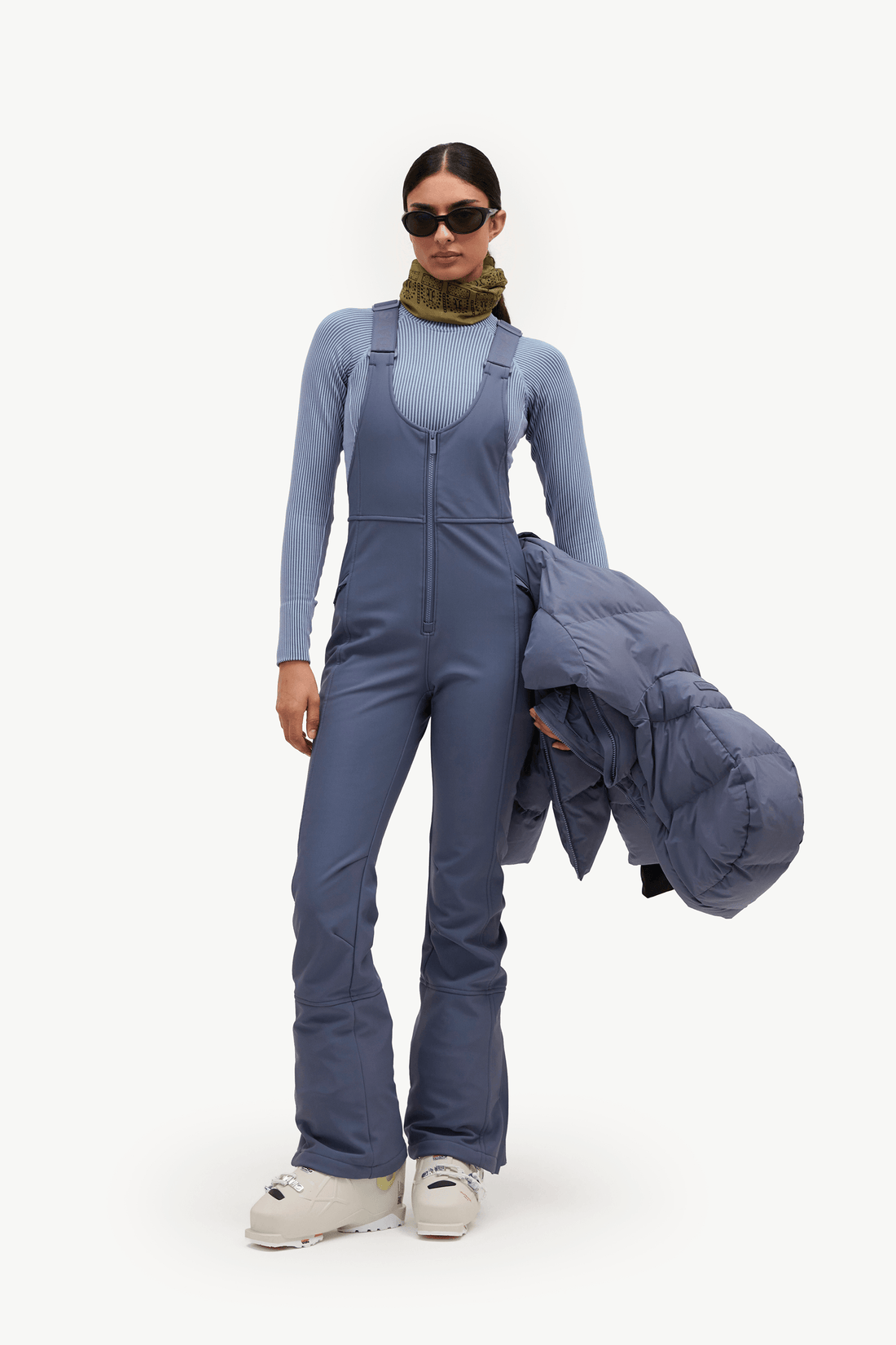 A model stands against a plain white studio background wearing slate blue ski bib overalls over a matching long-sleeve top, holding a matching slate blue puffer jacket, and wearing white sneakers.