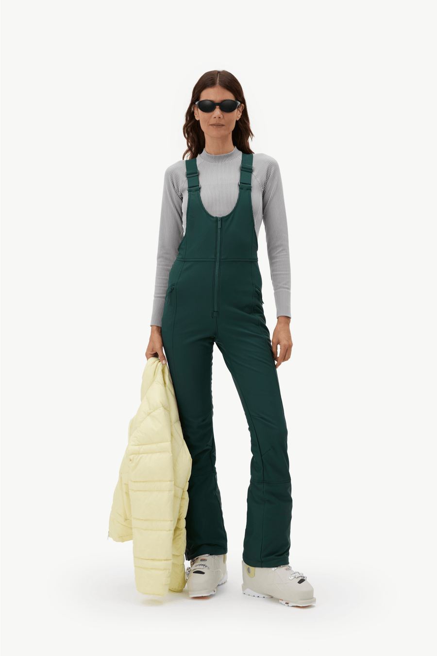 A model stands against a plain white studio background wearing dark green ski bib overalls over a light gray long-sleeve top, with white ski boots and sunglasses, and holding a pale yellow puffer jacket.