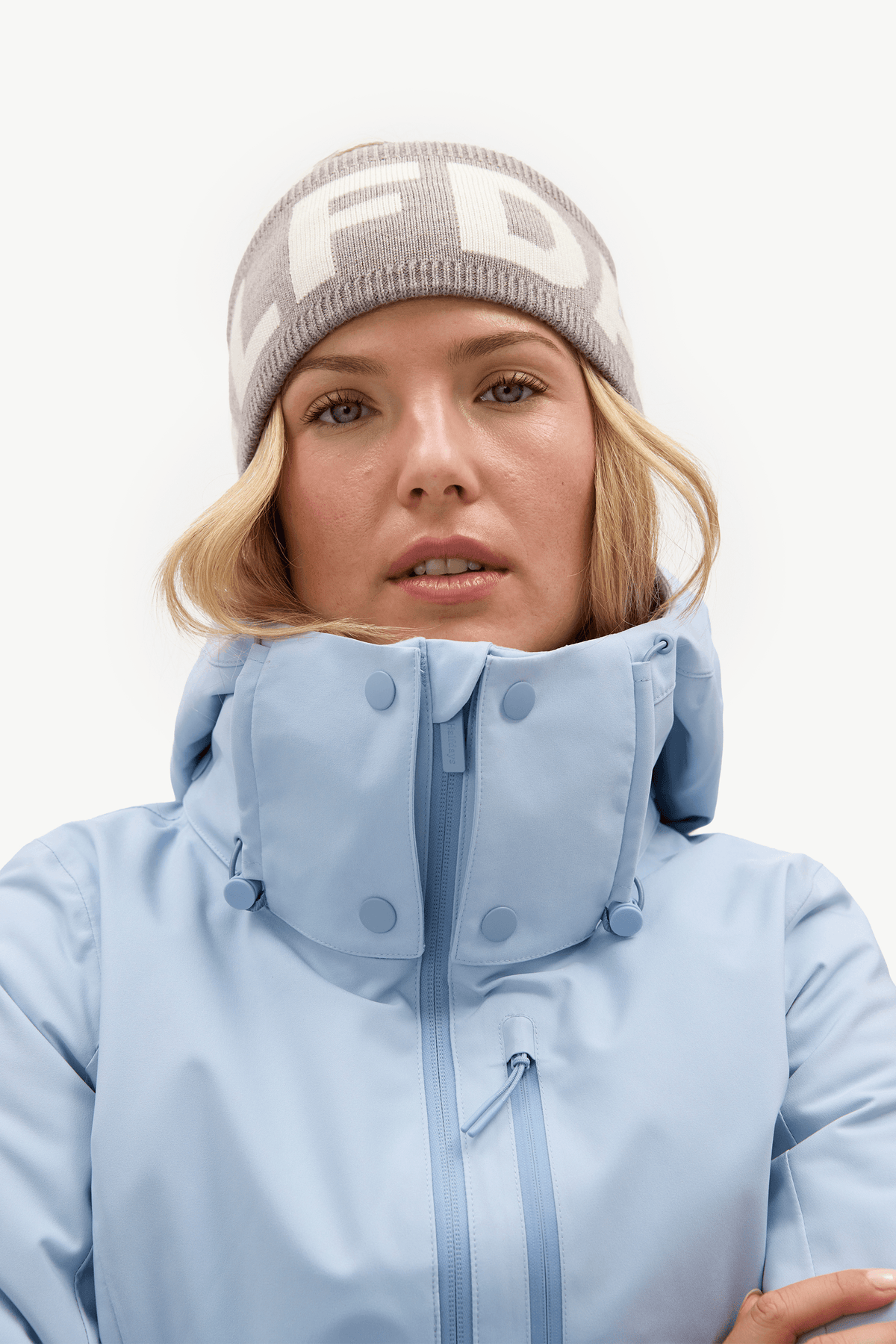 A woman wearing a light blue ski jacket with a high buttoned collar and a gray knit beanie with white lettering, photographed against a clean white studio background.
