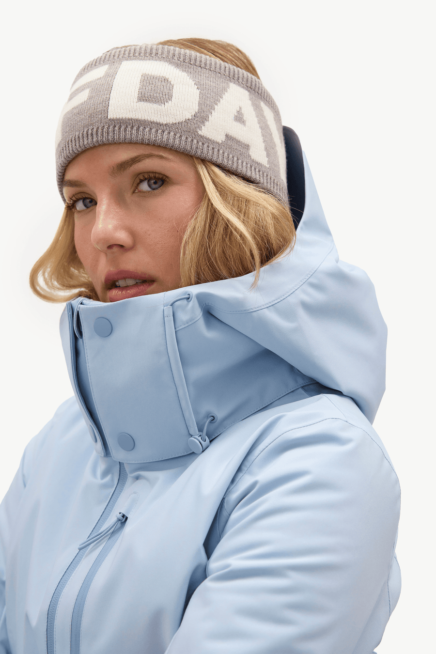 A close-up of a woman wearing a light blue ski jacket with a high collar and a gray knit beanie featuring the letters 'EDAI'.