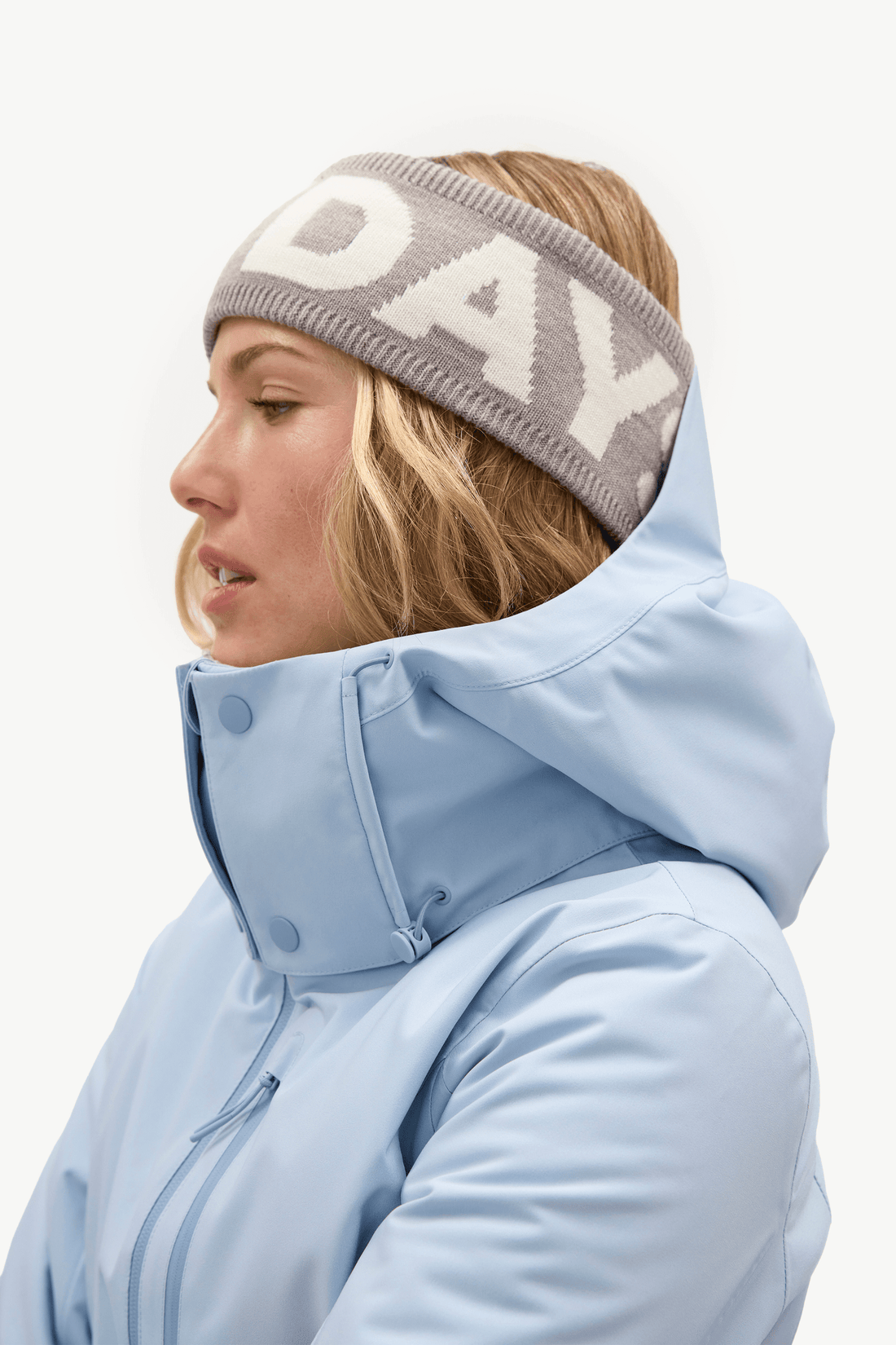 A woman in side profile wearing a light blue hooded winter jacket and a gray knit beanie with white lettering that reads 'DAY'.