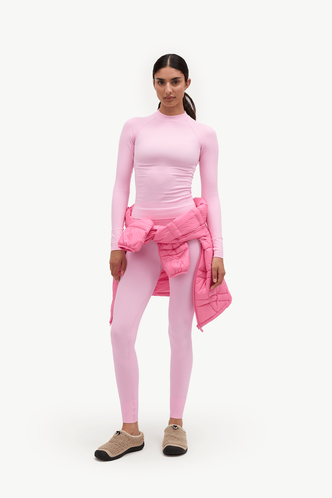 A model stands against a plain white studio background wearing a light pink long-sleeve mock-neck top, matching light pink leggings, and holding a pink puffer jacket tied around her waist. She is wearing beige slip-on shoes.