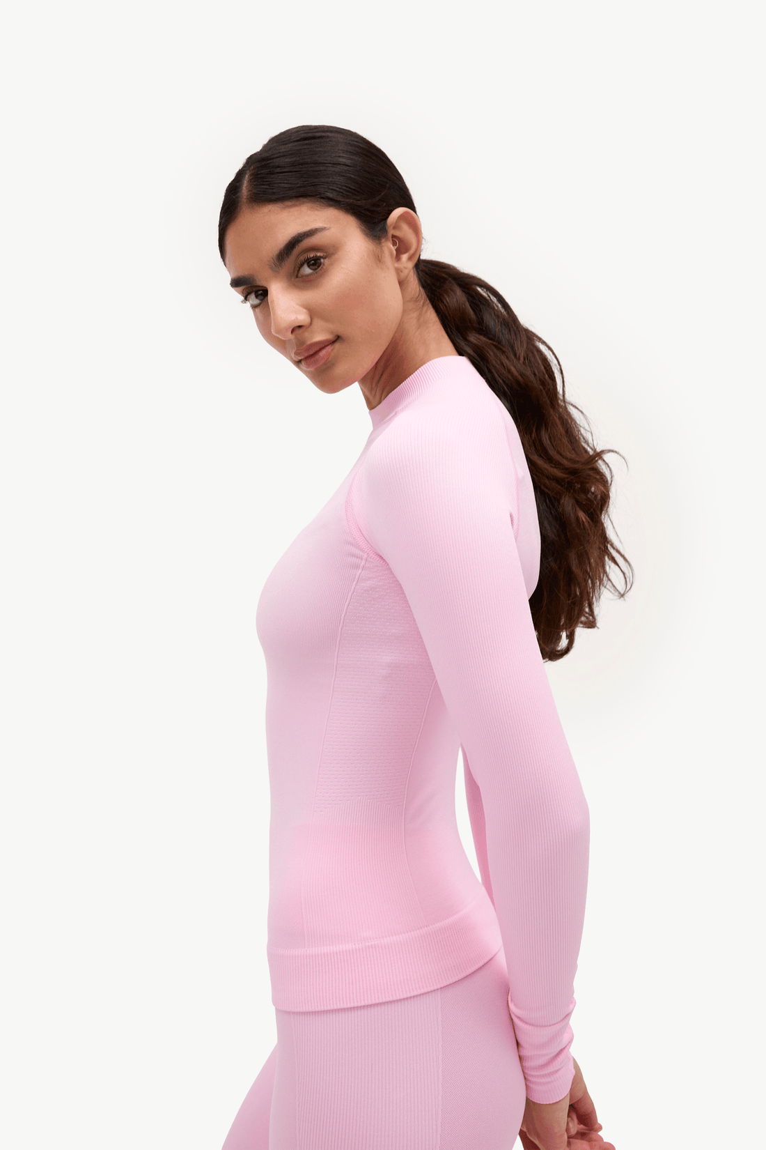 A woman with dark hair in a ponytail is shown from the side/back wearing a light pink long-sleeve athletic top and matching leggings against a plain white studio background.