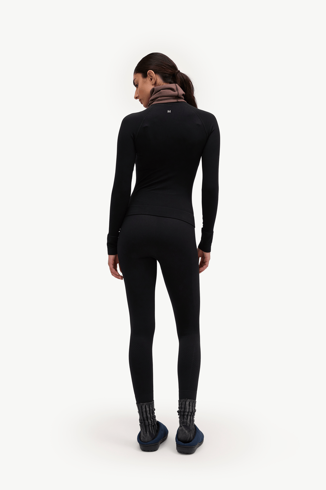 A woman is shown from behind wearing a black long-sleeve athletic top and matching black leggings, with a brown neck gaiter and dark blue shoes, against a plain white studio background.