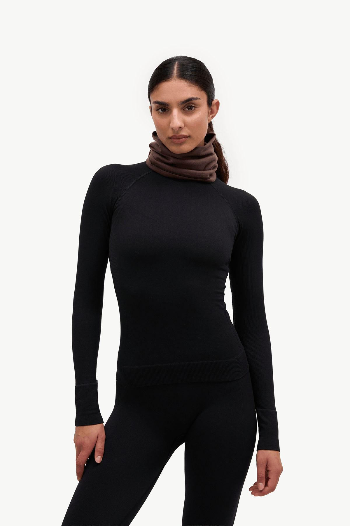 A woman modeling a black long-sleeve athletic top with a brown cowl neck scarf against a plain white studio background.