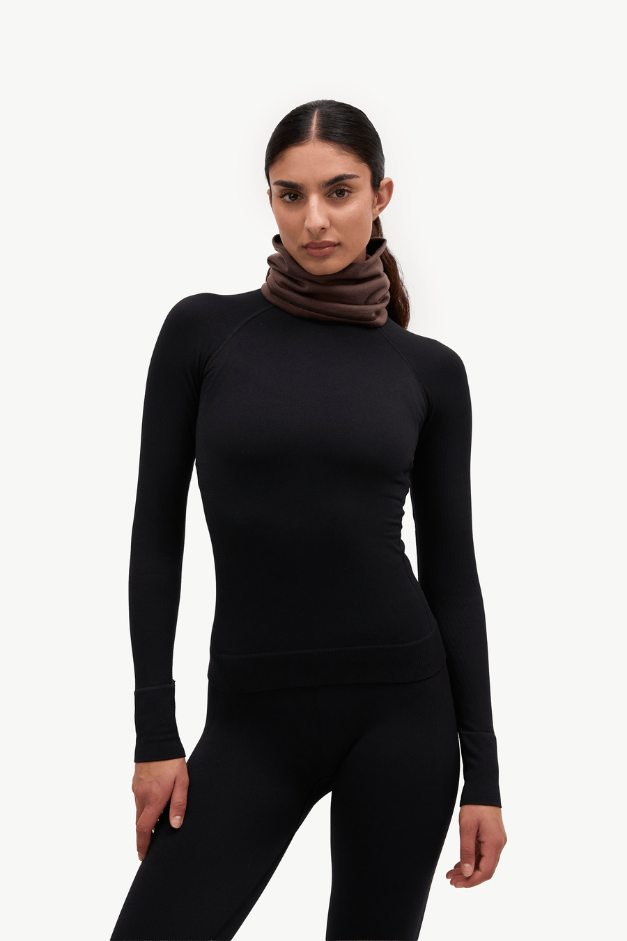 A woman modeling a black long-sleeve athletic top with a brown cowl neck scarf against a plain white studio background.