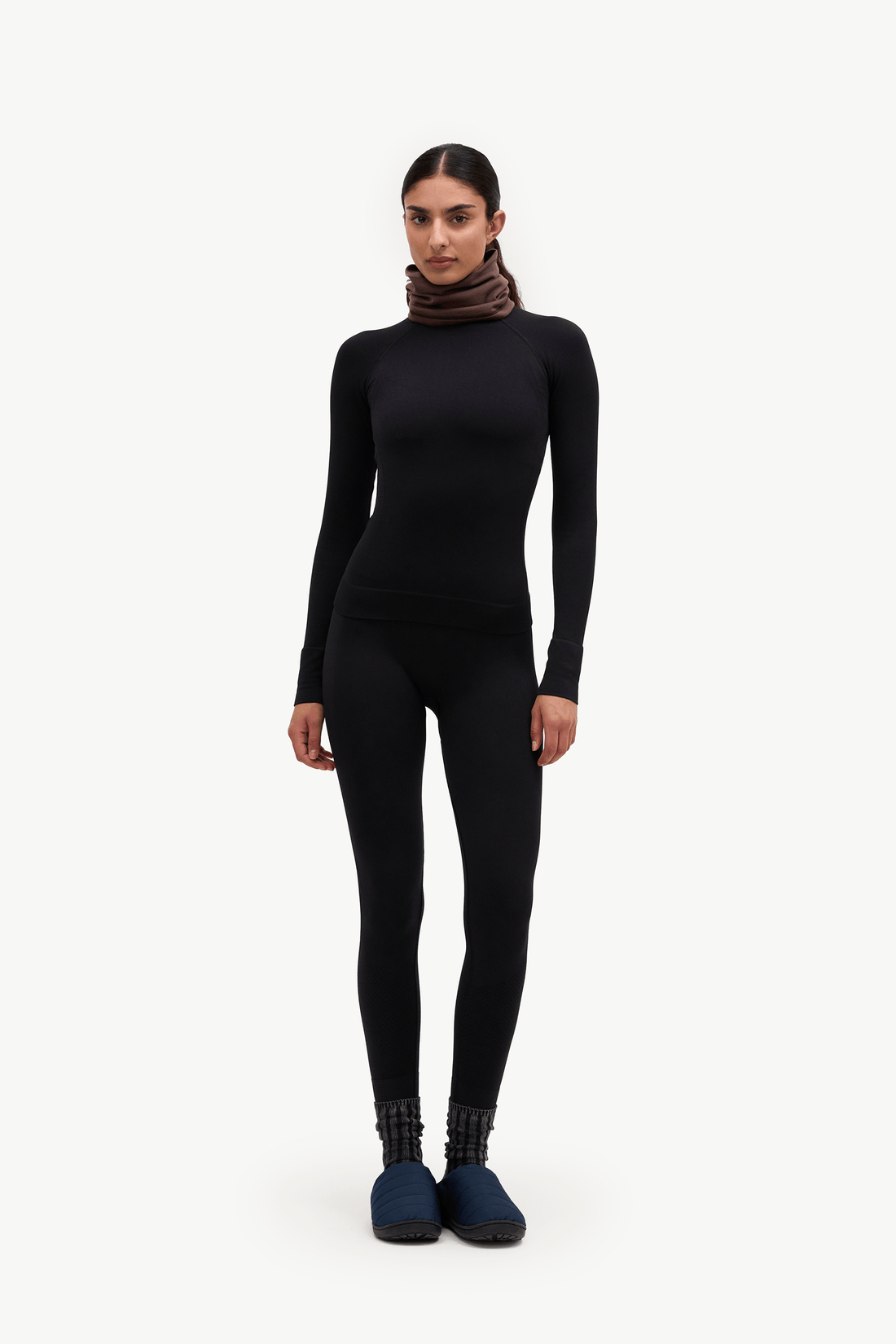 A model stands against a plain white studio background wearing a black long-sleeve turtleneck bodysuit, a dark brown knit neck warmer, and dark blue slip-on shoes.