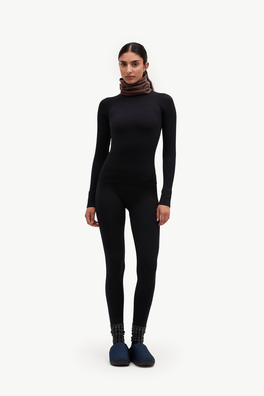 A model stands against a plain white studio background wearing a black long-sleeve turtleneck bodysuit, a dark brown knit neck warmer, and dark blue slip-on shoes.