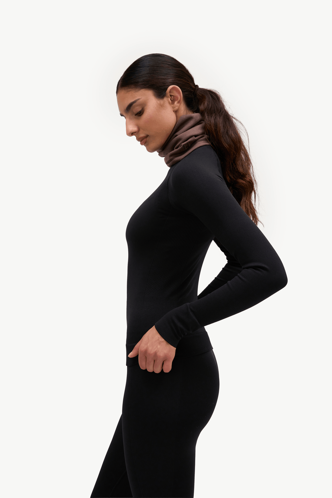A woman in a black long-sleeve bodycon turtleneck dress or bodysuit, shown in profile against a plain light gray studio background.
